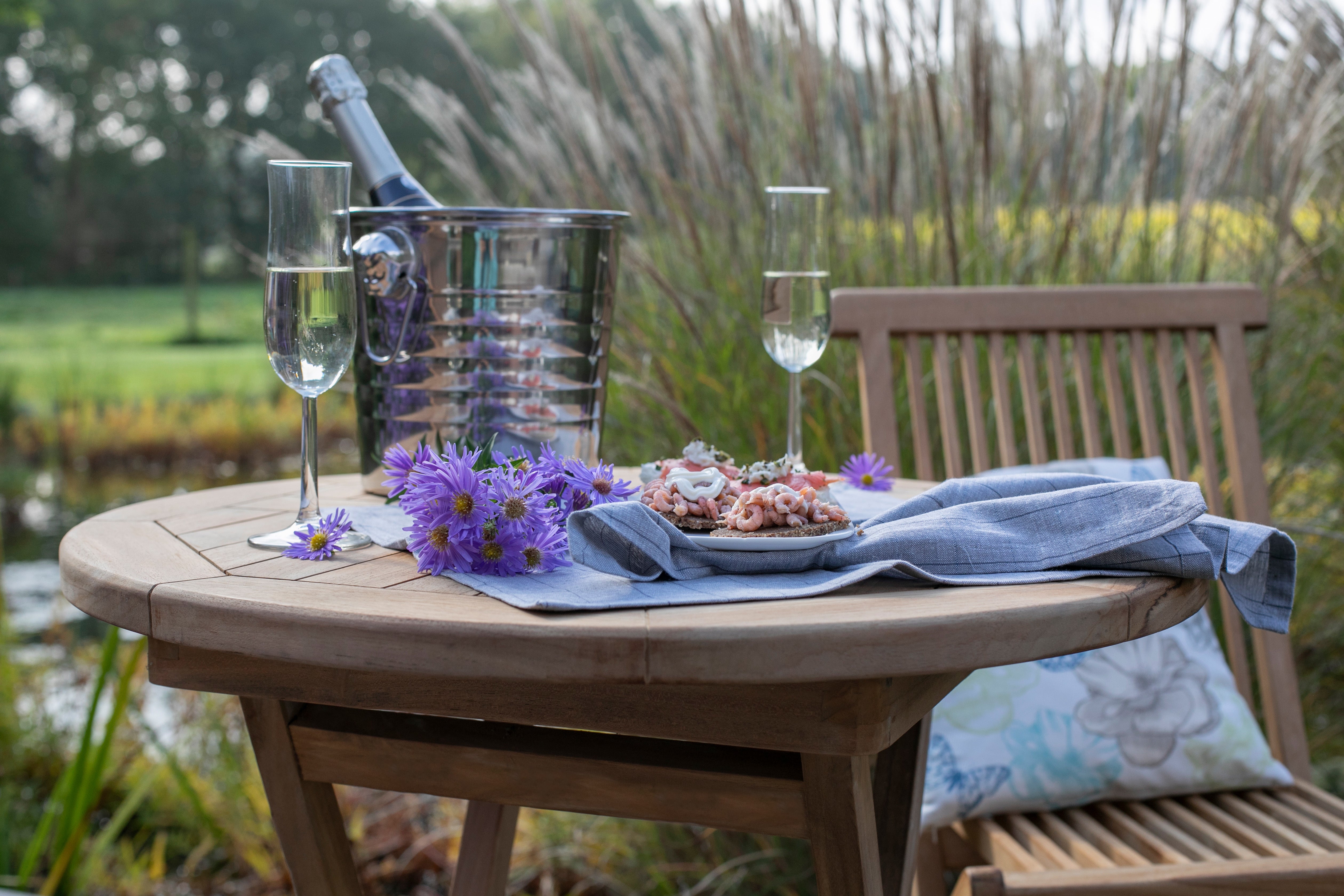 Auf einem Holztisch im Freien stehen zwei Gläser Champagner, köstliches Essen, lila Blumen und ein Strandkorb-Getränkekühler – ein elegantes Luxusaccessoire, das Ihre Flasche bei jeder Feier gekühlt und wetterfest hält.