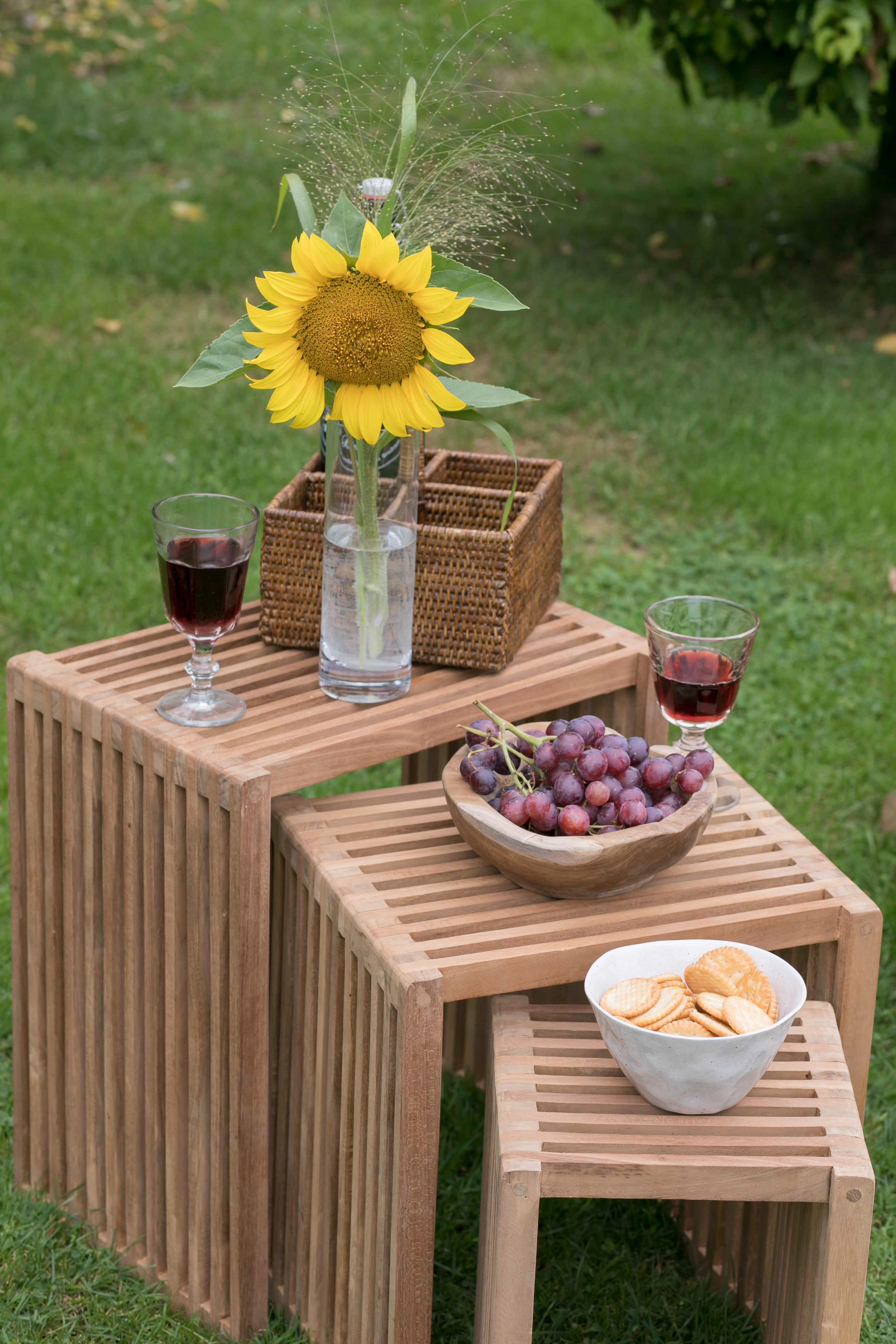 Das Beistelltische 3er-Set wird im Freien auf einer Wiese mit Wein, Weintrauben, Crackern und einer Vase mit einer Sonnenblume arrangiert.