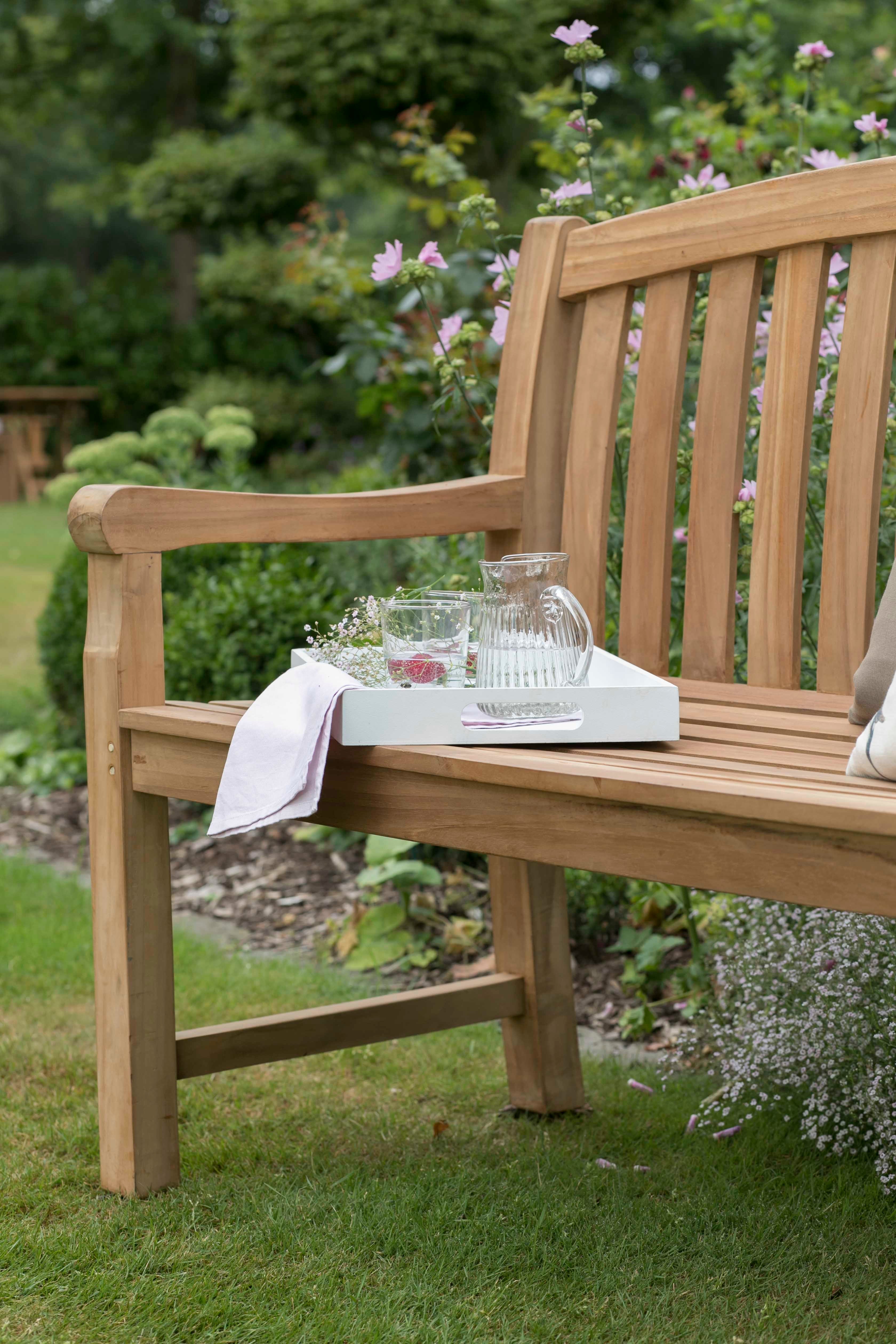 Gartenbank Swindon aus Teakholz mit ergonomischer Form, Ablage für Glas, Kanne und Tuch, umgeben von Blumen.