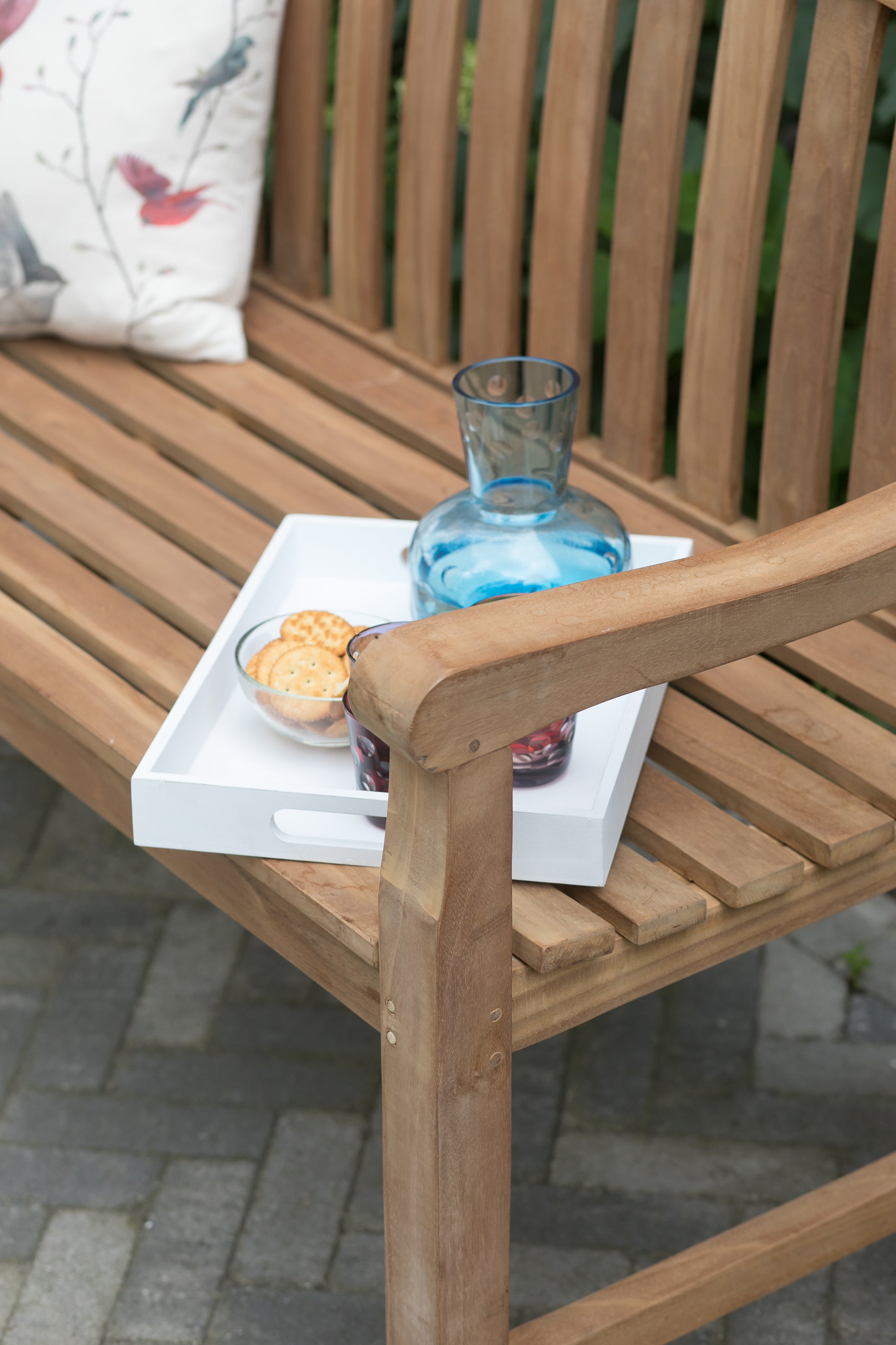Auf der Gartenbank Swindon mit ergonomischer Formgebung stehen ein Tablett mit Glaskaraffe, Gläsern und Keksen – alles aus edlem Naturholz – im Freien.