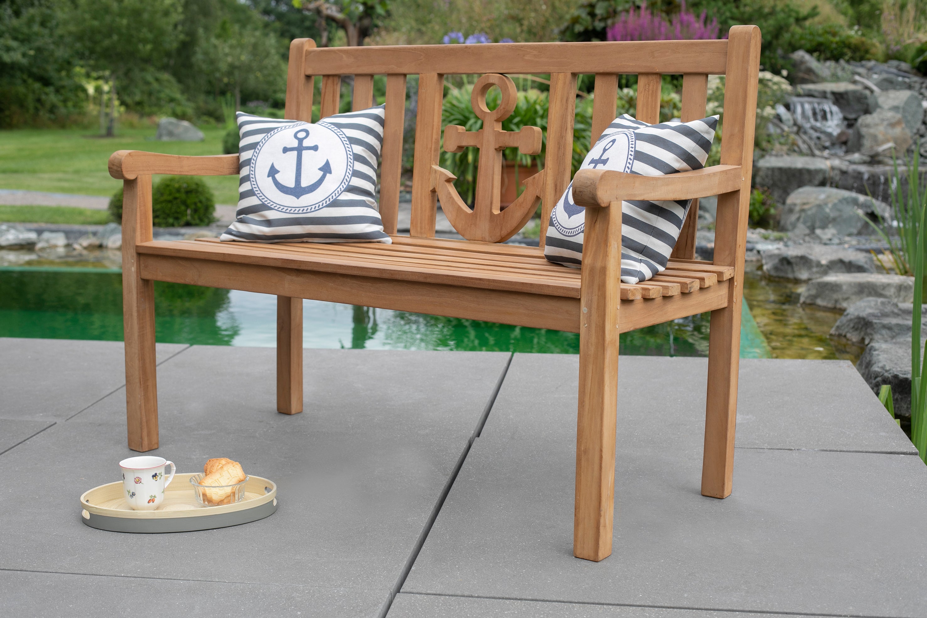 Die Teak-Gartenbank Anker aus massivem Naturholz mit passendem Kissen lädt am Teich oder auf dem gefliesten Patio zu entspannten Momenten ein – stilvolle Outdoor-Möbel für Genießer.