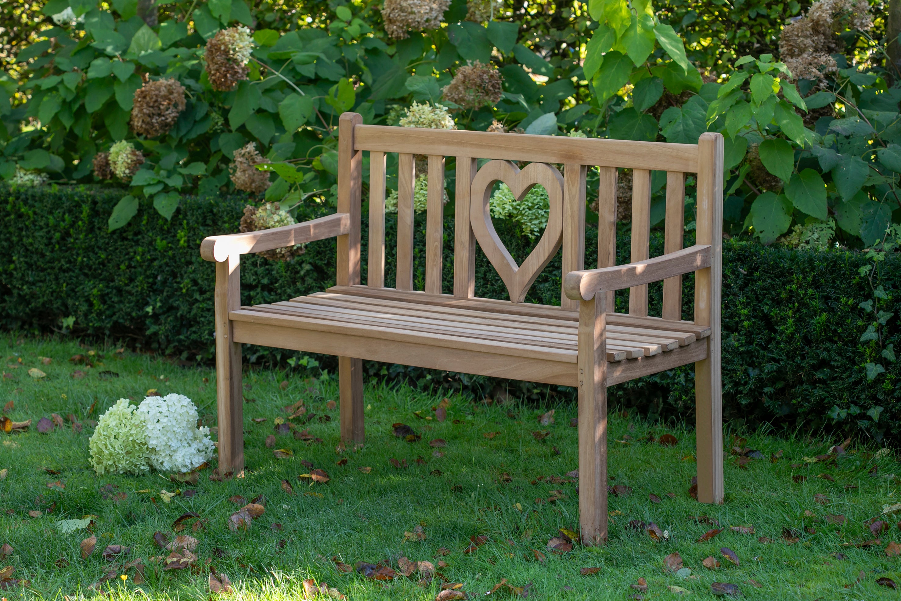 Die aus natürlichem Massivholz gefertigte Teak-Bank Motivbänke mit herzförmigem Rückenausschnitt verleiht Außenbereichen Charme und sieht auf Gras in der Nähe von grünen Büschen perfekt aus.