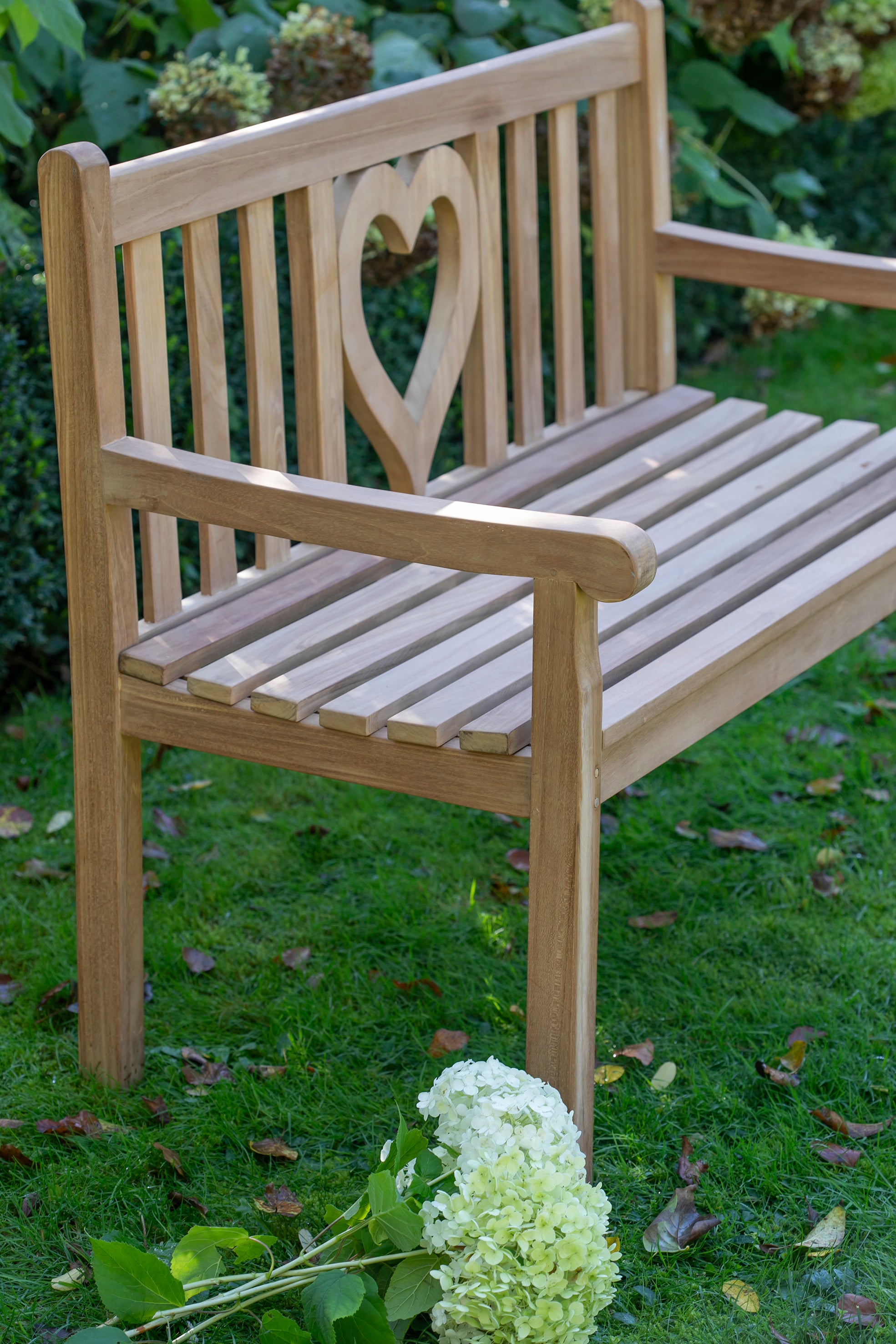 Die Motivbänke Teak mit herzförmigem Ausschnitt an der Rückenlehne ist ein einladendes Gartenmöbelstück, das neben einem Strauß weißer Blumen auf der Wiese platziert wird.