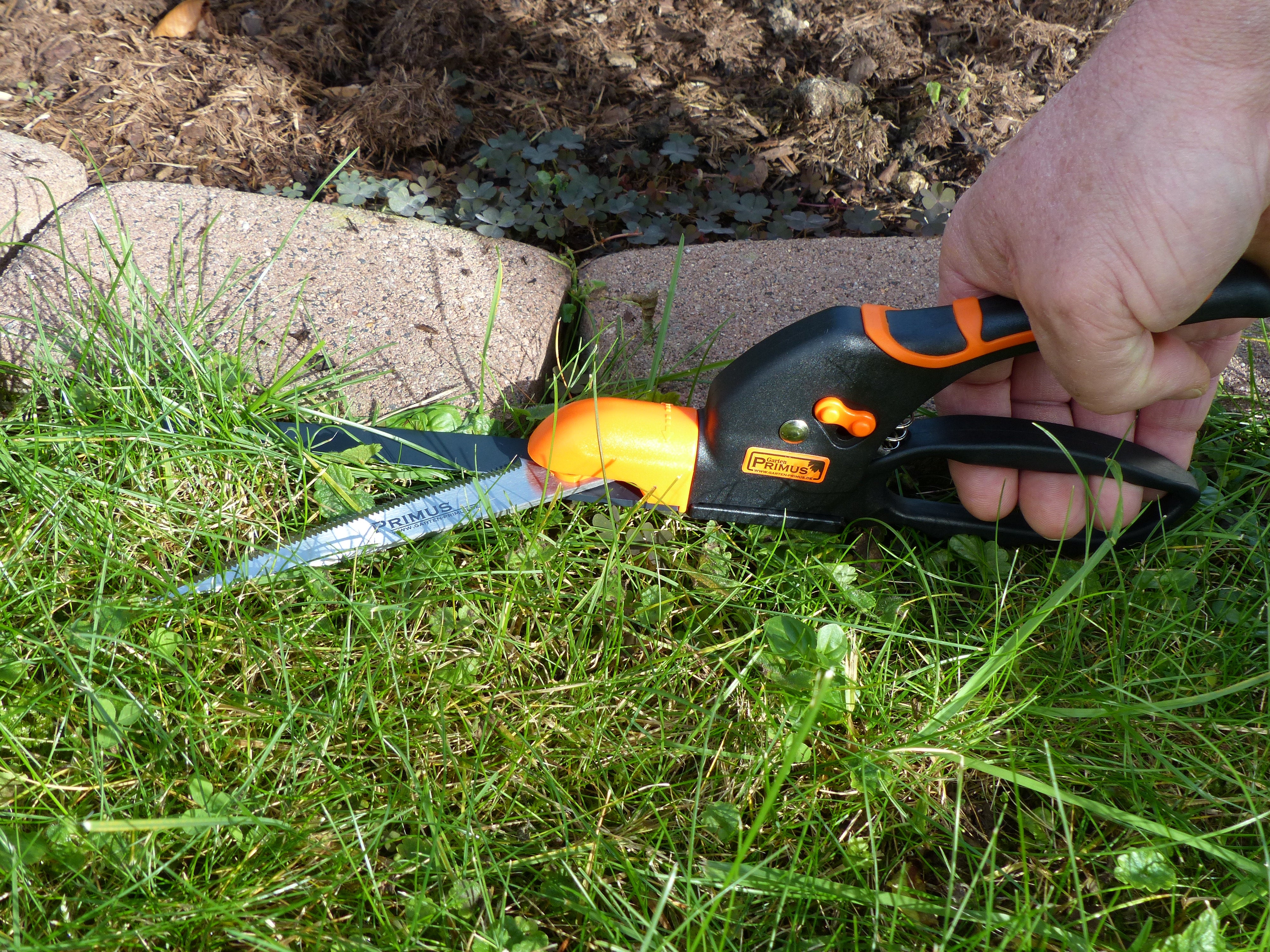 Mit der Rasenkanten-Schere schneidet eine Hand die Rasenkante an einem mit Steinen eingefassten Gartenbeet – das Multifunktionswerkzeug garantiert stets saubere und präzise Ergebnisse.