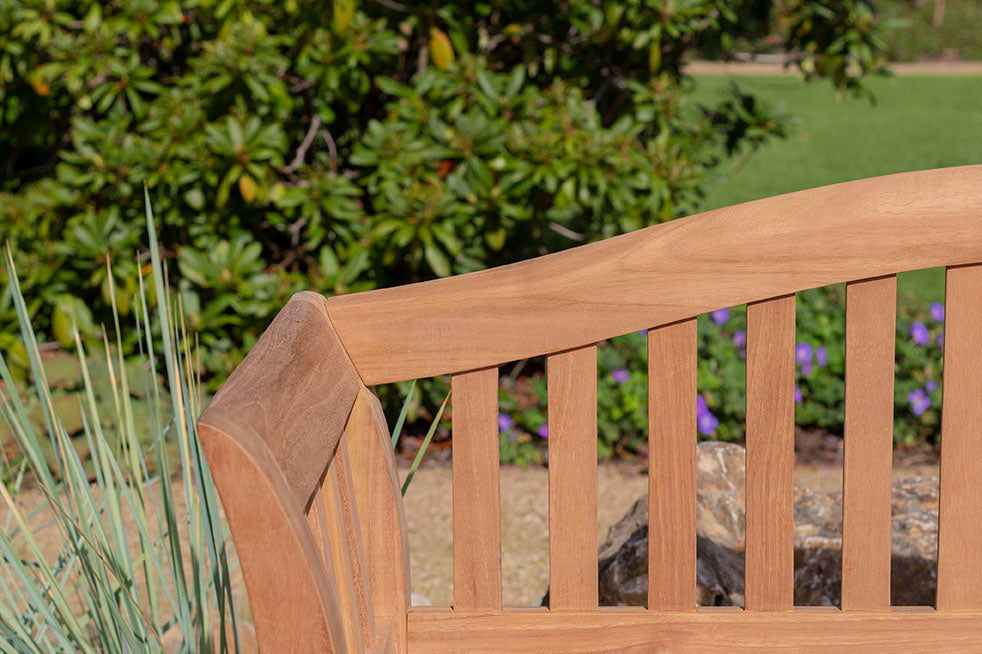 Nahaufnahme der Gartenbank Wave mit vertikalen Lamellen aus Massivholz, in einem Garten umgeben von Grün und lila Blumen – das perfekte Outdoor-Möbel für entspannende Momente.