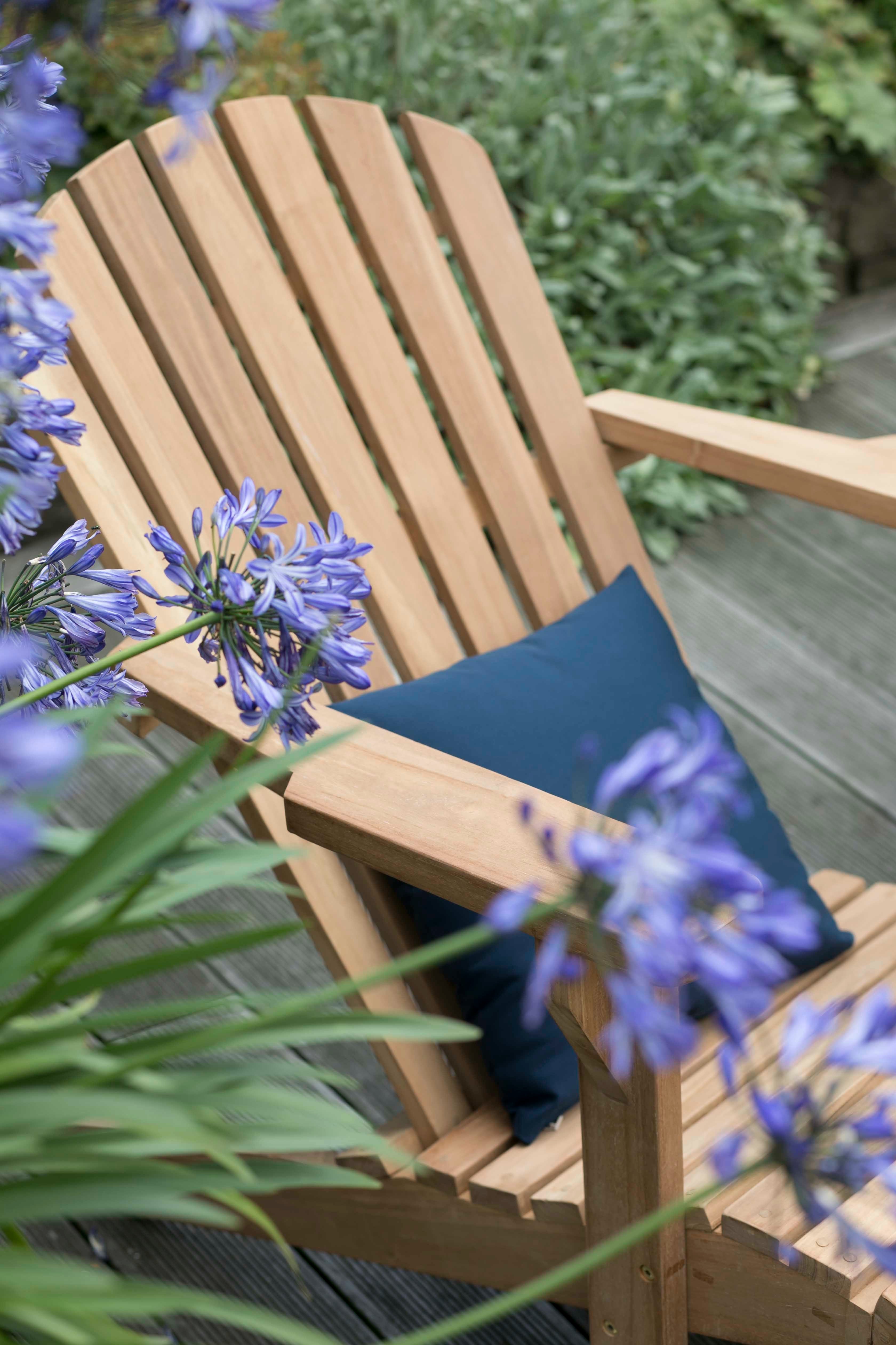 Langlebige Qualität: Der Gartenlounger Adirondack Montreal mit blauen Kissen ist witterungsbeständig und steht zwischen grünen Pflanzen sowie violetten Blumen.