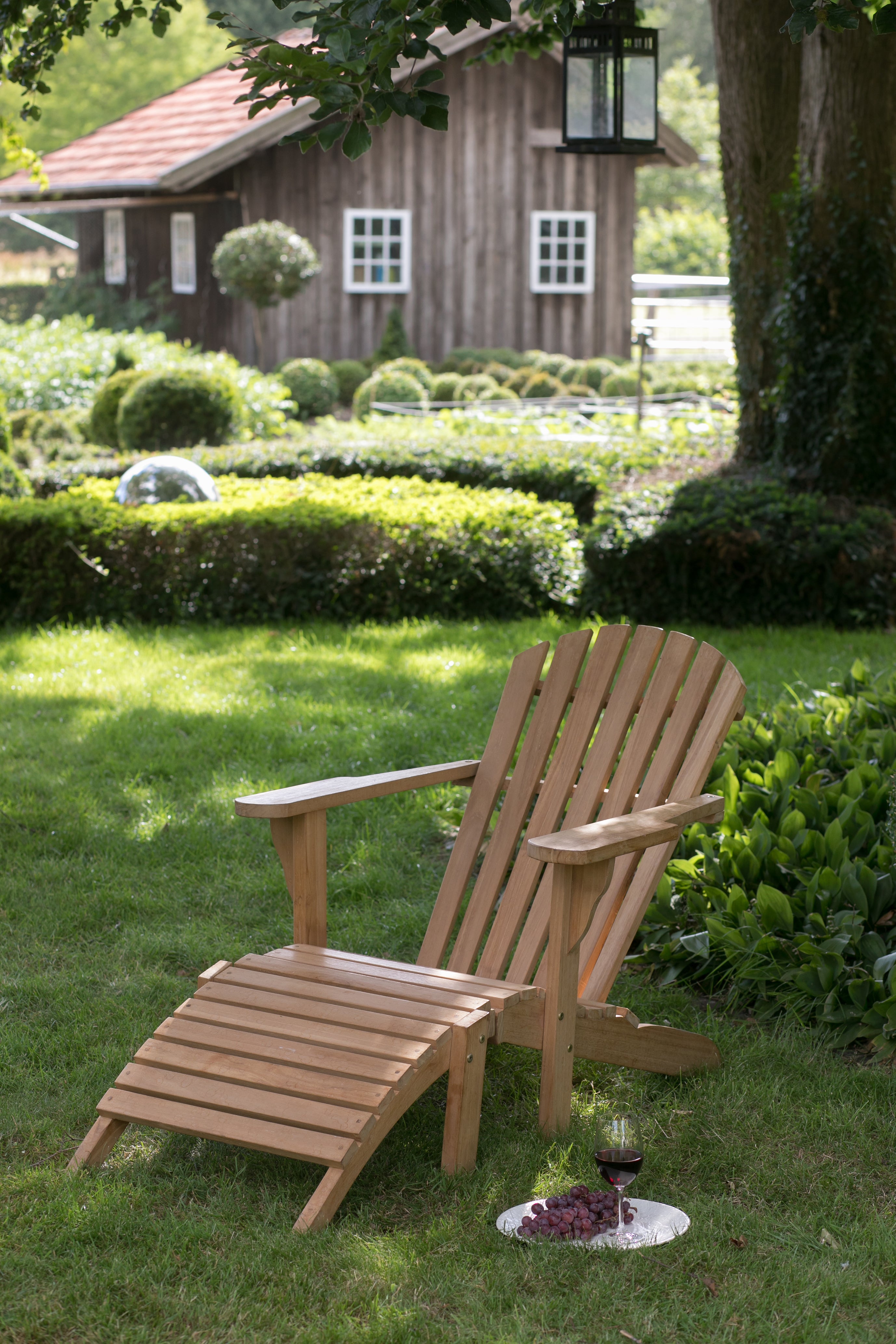 Der Gartenlounger Adirondack Montreal mit passendem Ottoman steht wetterfest auf dem Rasen, neben Trauben und Wein; Im Hintergrund ist ein gemütliches Cottage zu sehen.