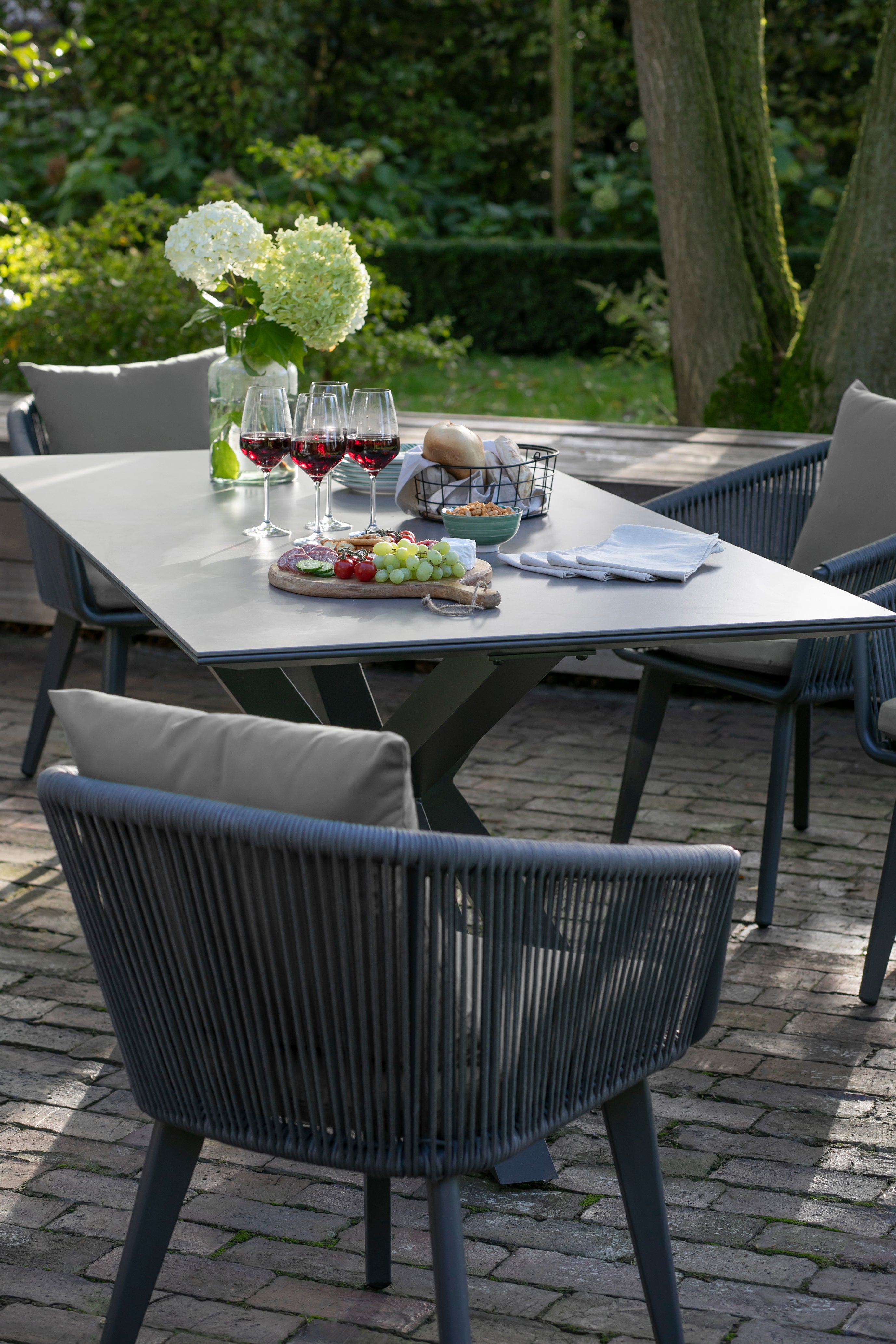 Das Gartenmöbelset Rope & Alu, bestehend aus Weingläsern, Käse, Trauben, Brot und Blumen, ist von stilvollen Stühlen umgeben und sorgt für ein perfektes, modernes Esserlebnis im Freien.