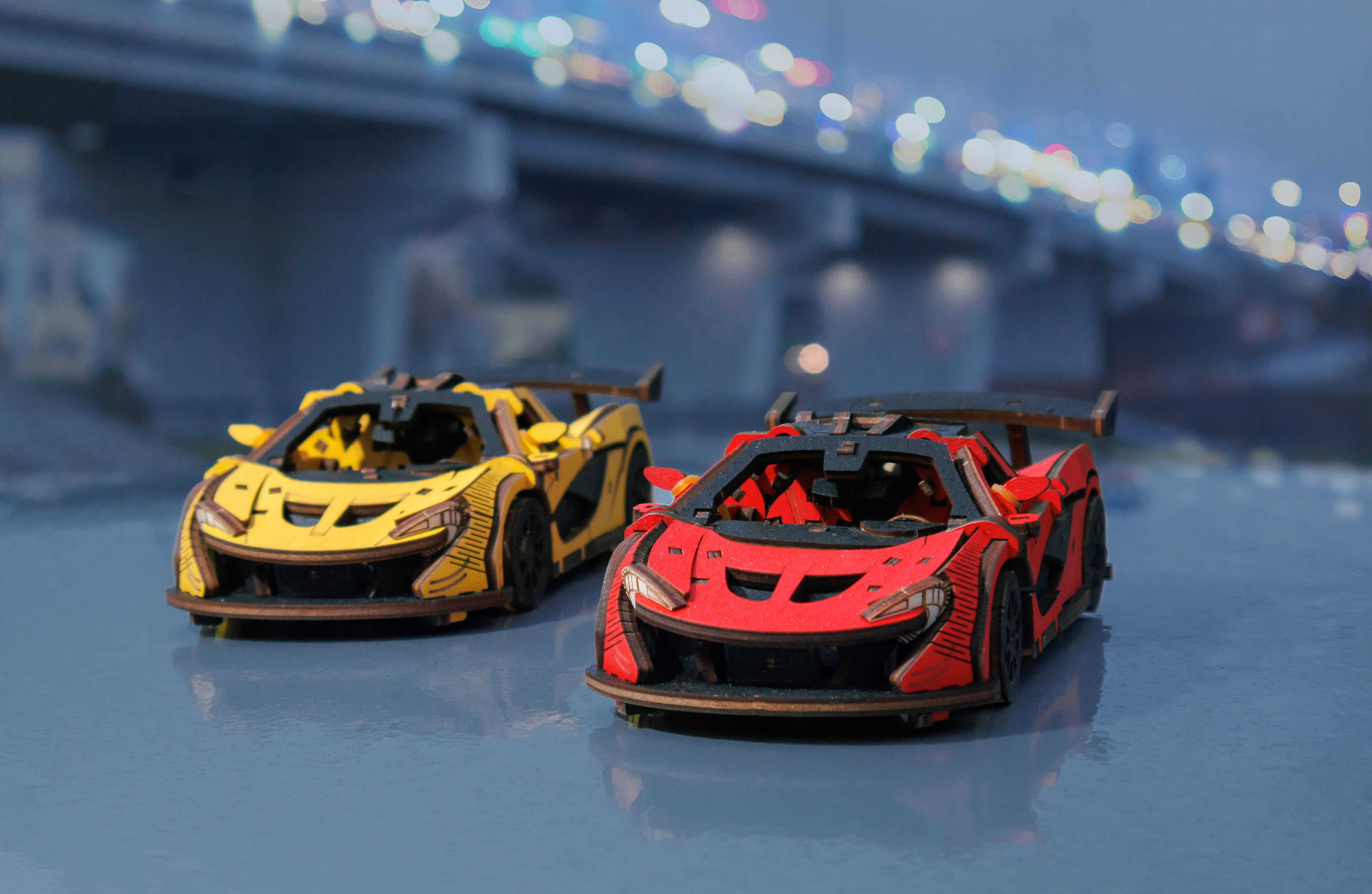 Das 3D-Holzpuzzle Racing Car gelb steht auf einer glänzenden Oberfläche vor unscharfem Brückenhintergrund – ein Muss für Rennsportbegeisterte und Modellbau-Fans.