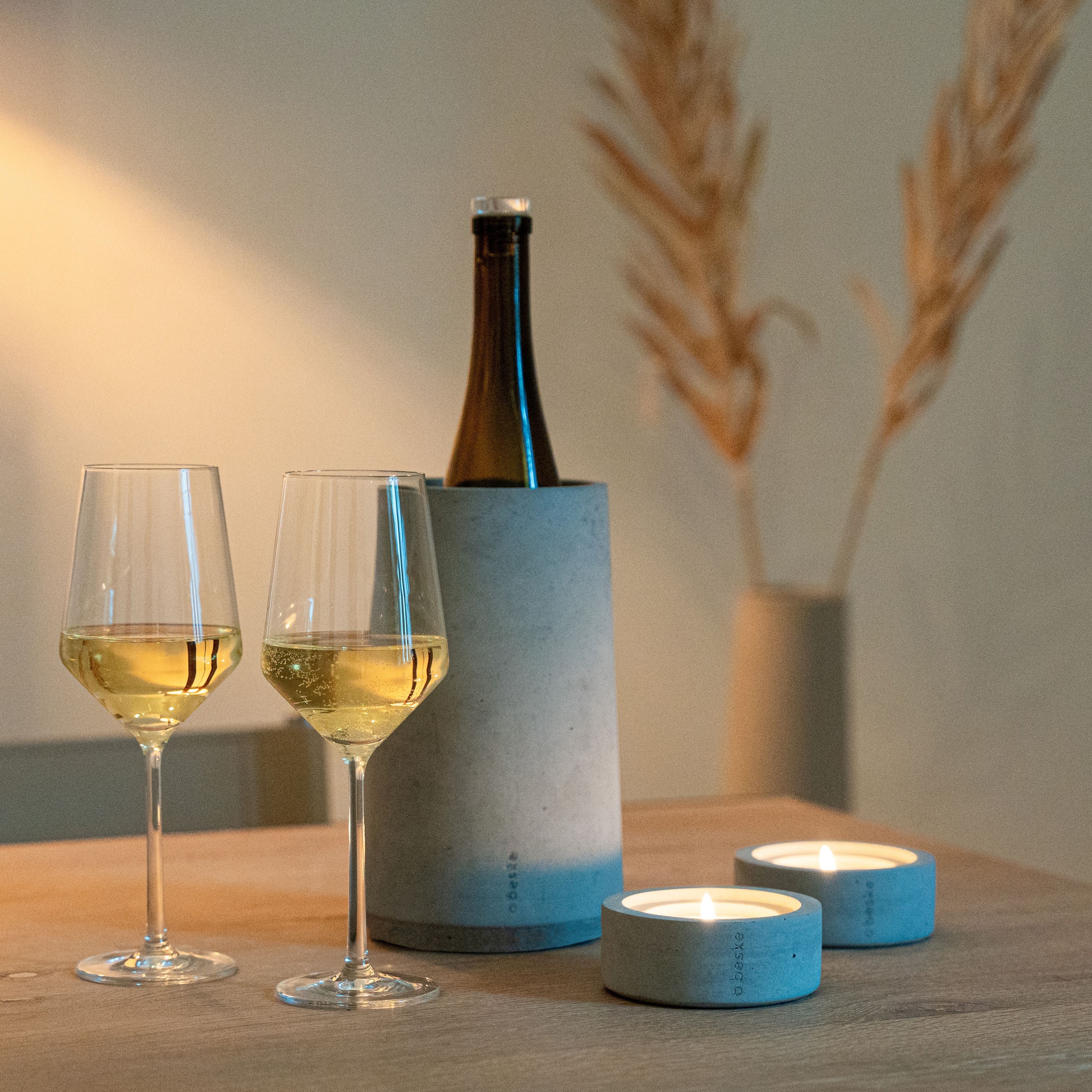 Zwei Gläser Weißwein, eine Weinflasche im Beske Weinkühler „Eiskar“ und zwei brennende Kerzen stehen auf einem Holztisch.