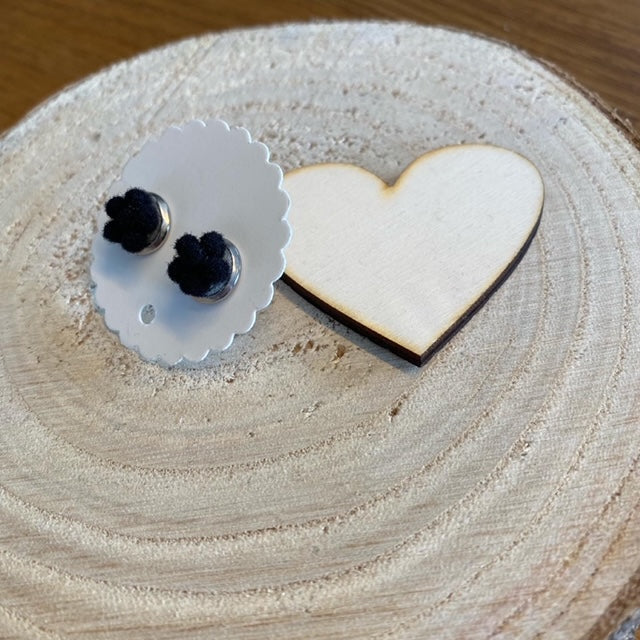 Eine Ohrringkarte mit Wellenmuster präsentiert Bollenhut-Ohrringe neben einem Holzherz auf einer runden, hellen Holzoberfläche und unterstreicht den Charme der Schwarzwälder Handwerkskunst.