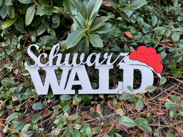 Ein Schwarzwald-Schriftzug mit rotem Hut-Akzent, inspiriert vom Bollenhut-Design, liegt im Freien auf grünem Boden.