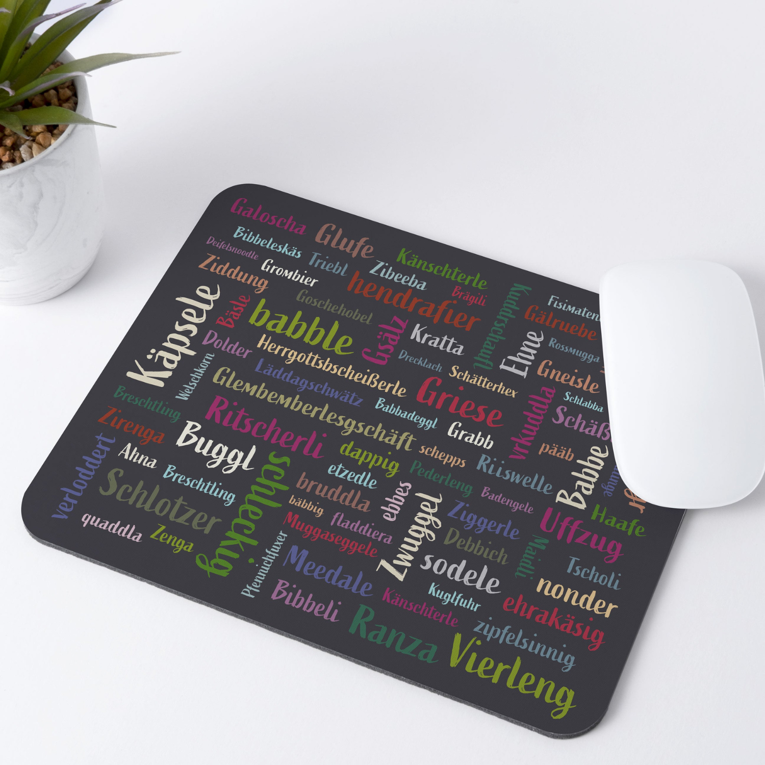 Eine weiße Maus und das Mundart-Mousepad mit bunten deutschen Wörtern im Mundart-Design liegen neben einer kleinen Topfpflanze auf einem weißen Schreibtisch, sorgen für Flair und schützen die Oberfläche.