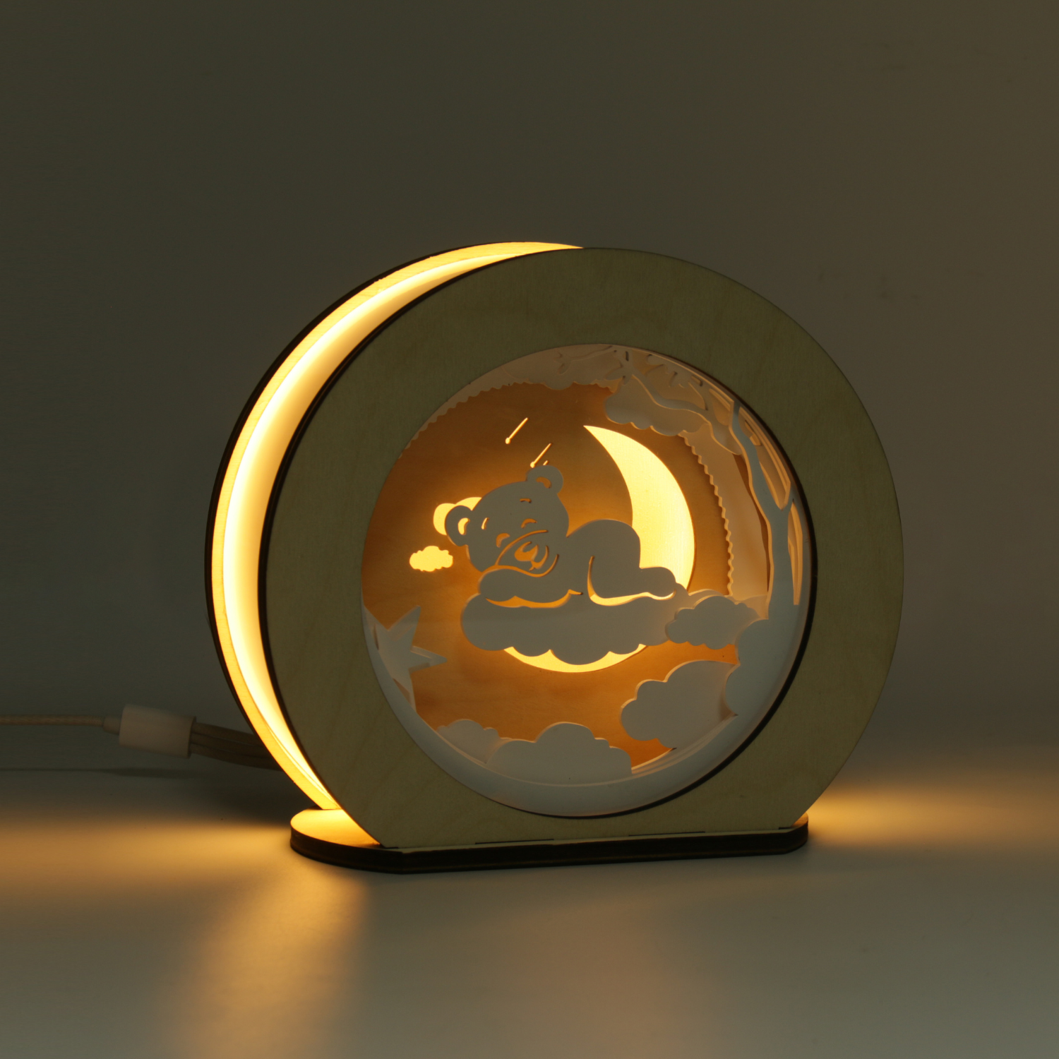 Die Magic Light 3D Holz-Nachtlampe Wolkenbär ist eine runde Nachtlampe mit leuchtendem Mond, Wolken und Bär, die warmes LED-Licht für eine beruhigende Atmosphäre auf dunklem Hintergrund wirft.
