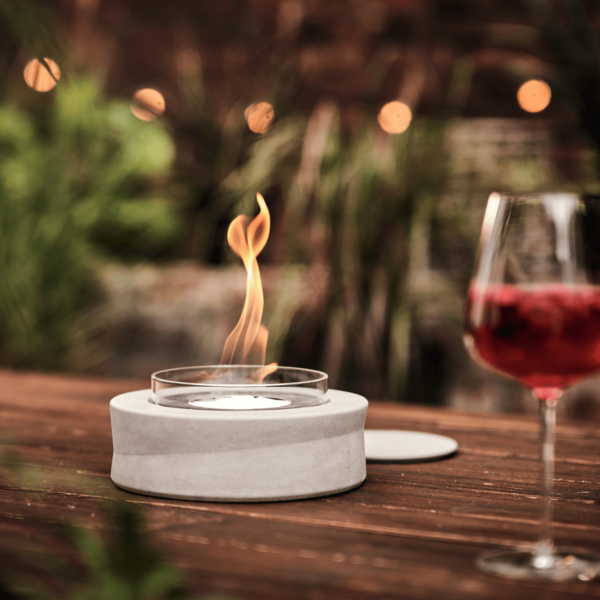Ein Ethanolfeuer der Beske Manufaktur brennt auf einem Holztisch neben einem Glas roten Getränks und sorgt für eine gemütliche Atmosphäre im Freien.