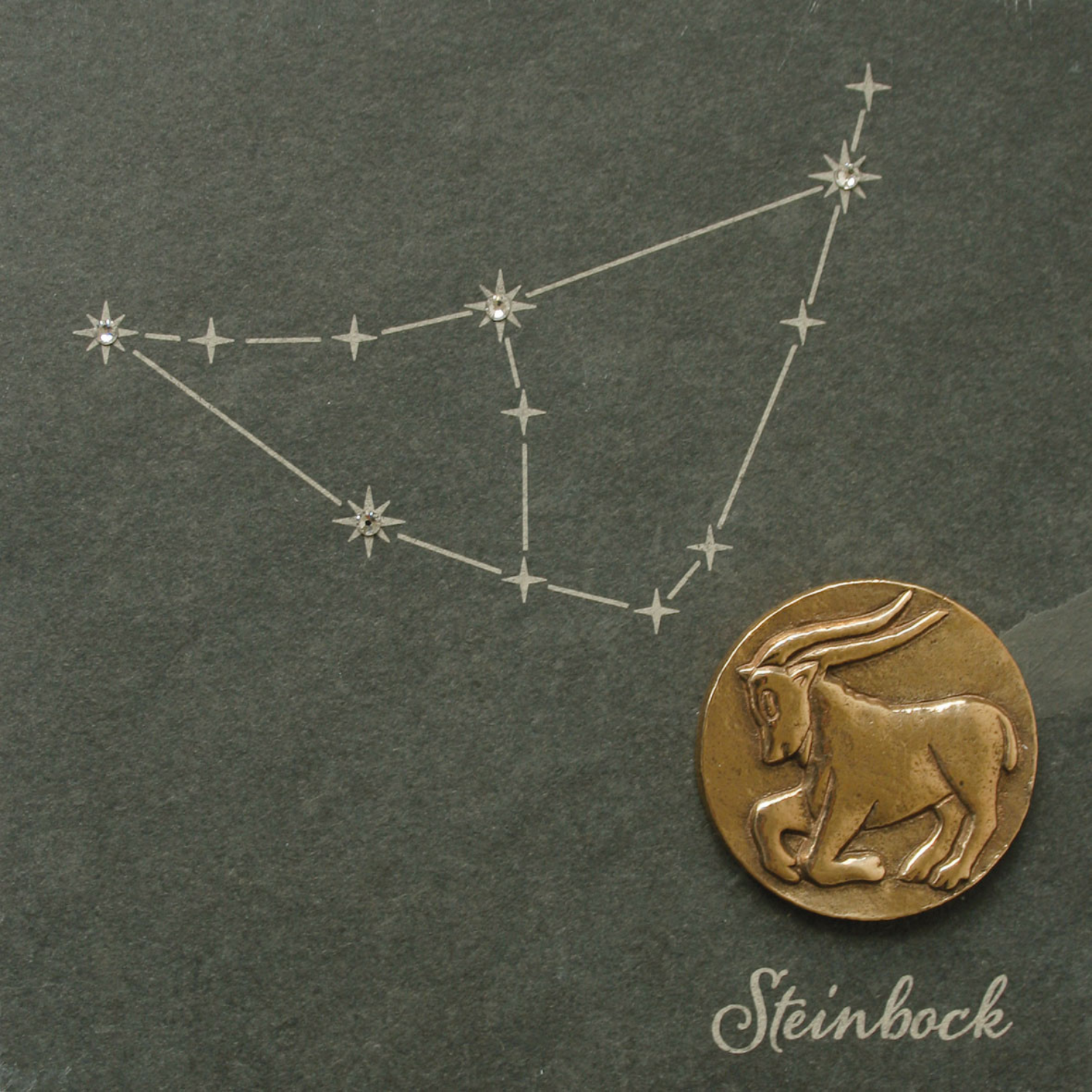 Eine Bronze-Sternzeichen-Münze mit Swarovski Elements ruht neben einer Zeichnung des Sternbildes Steinbock auf einem dunklen Naturschiefer-Hintergrund mit elegantem Steinbock-Text.