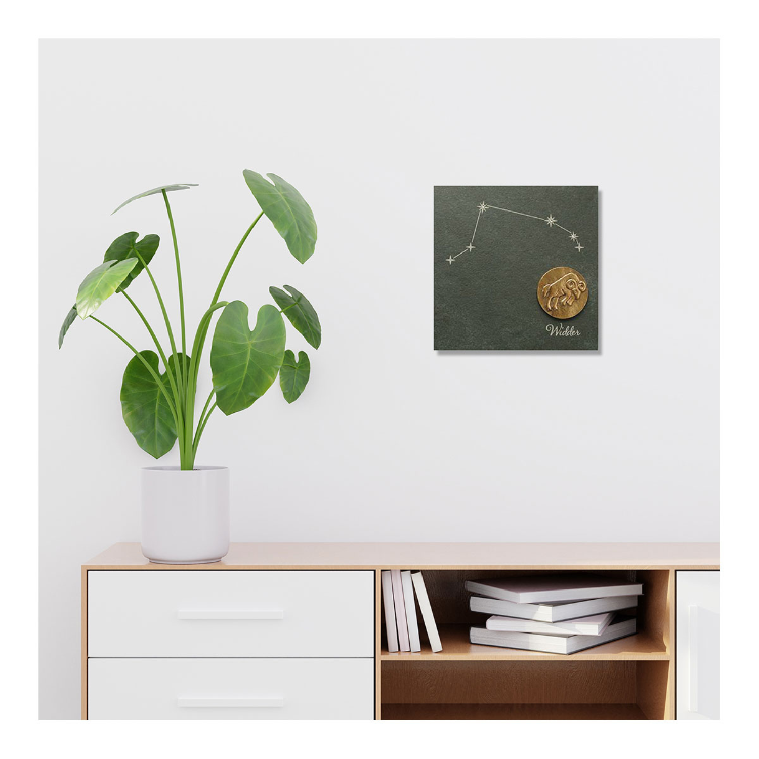 Auf einem weißen Schrank steht eine Topfpflanze, über der das Wandbild mit dem Sternzeichen Waage in Bronze hängt.
