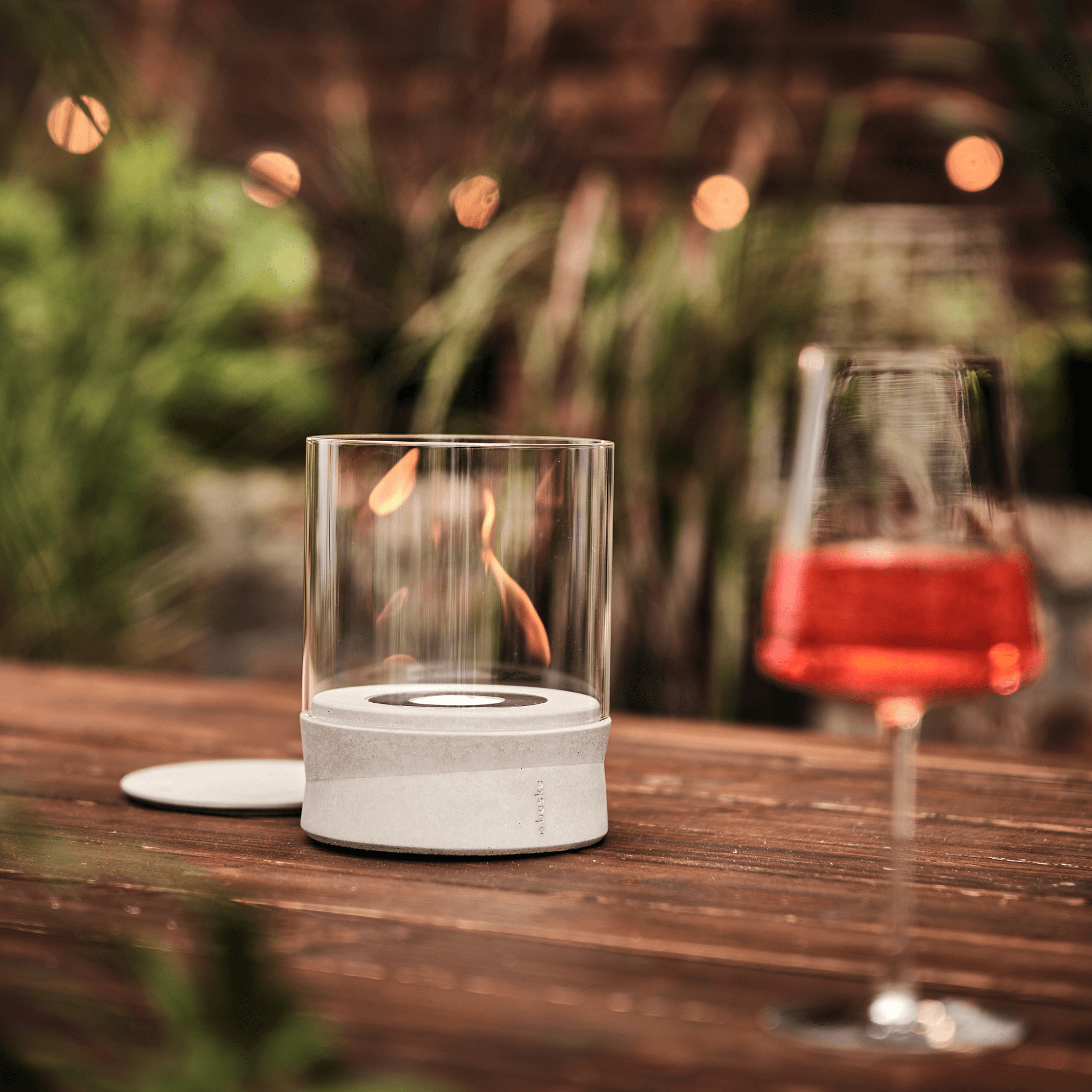Das Ethanolfeuer von Beske Manufaktur und ein Glas rotes Getränk stehen auf einem hölzernen Außentisch mit Pflanzen im Hintergrund.