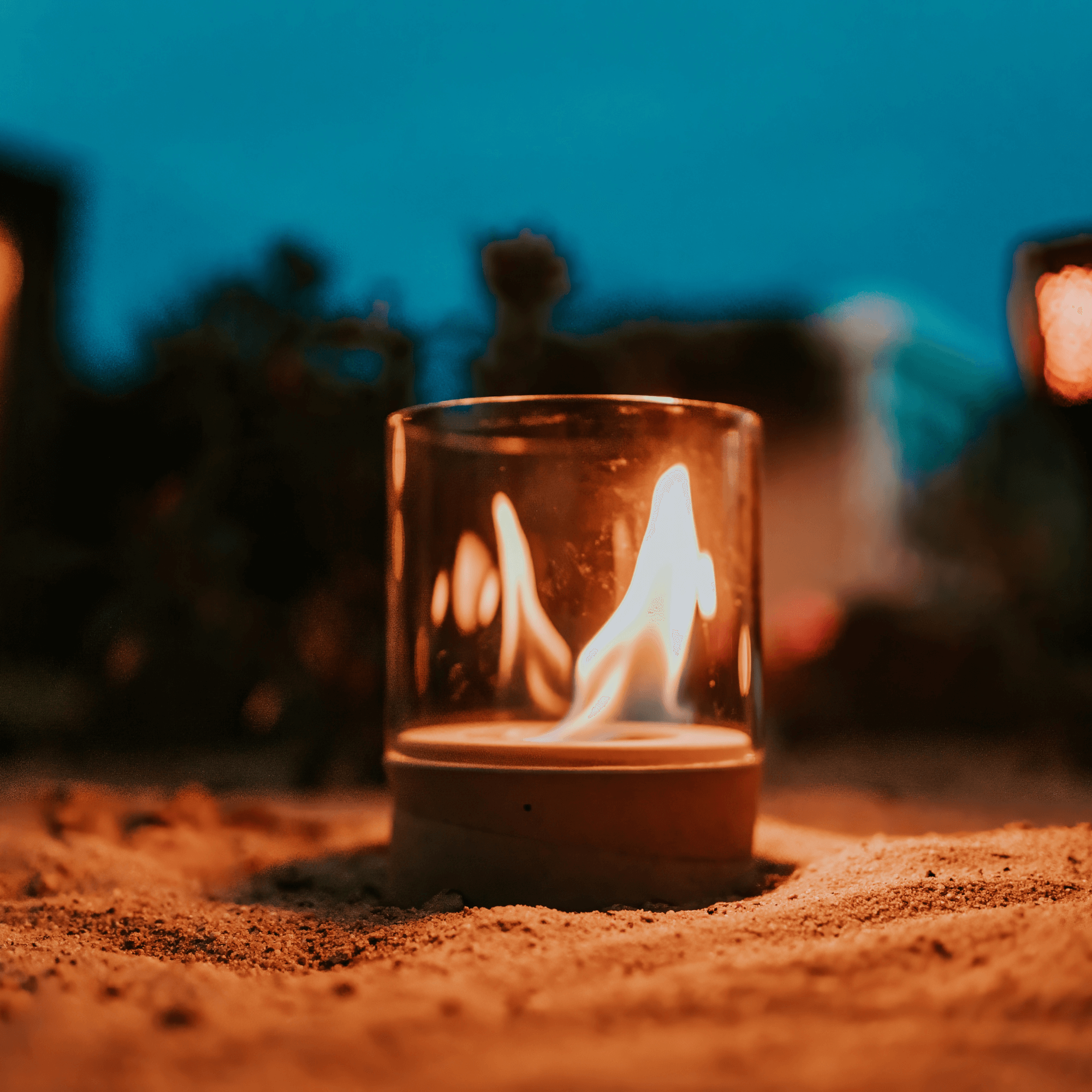 Das Ethanolfeuer der Beske Manufaktur, ein glasummanteltes Bioethanolfeuer, das an einen Tischkamin erinnert, steht in der Dämmerung glühend auf sandigem Boden.