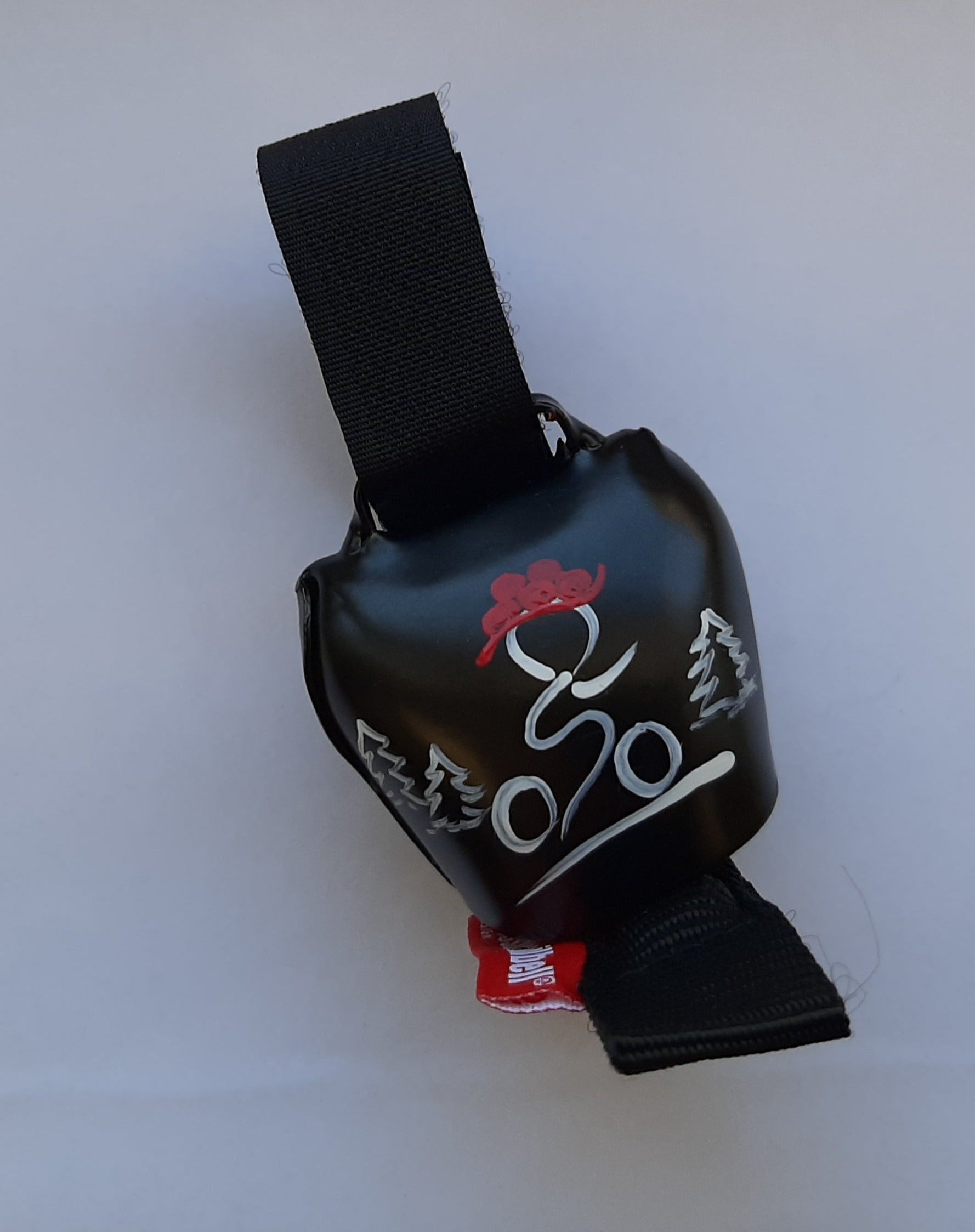 Eine schwarze Fahrradglocke swisstrailbell® mit Riemen, die einen Radfahrer als Strichmännchen mit rotem Helm zwischen zwei Bäumen auf weißem Hintergrund zeigt.