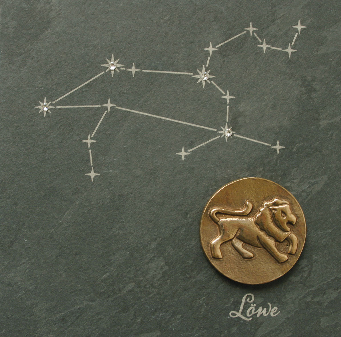 Eine bronzene Löwenmünze und das Sternbild Löwe schimmern auf dunklem Hintergrund mit dem Schriftzug „Löwe“, akzentuiert durch Swarovski Elements und auf einem eleganten Naturschiefer. Wir präsentieren die Bronze-Sternzeichen-Kollektion.