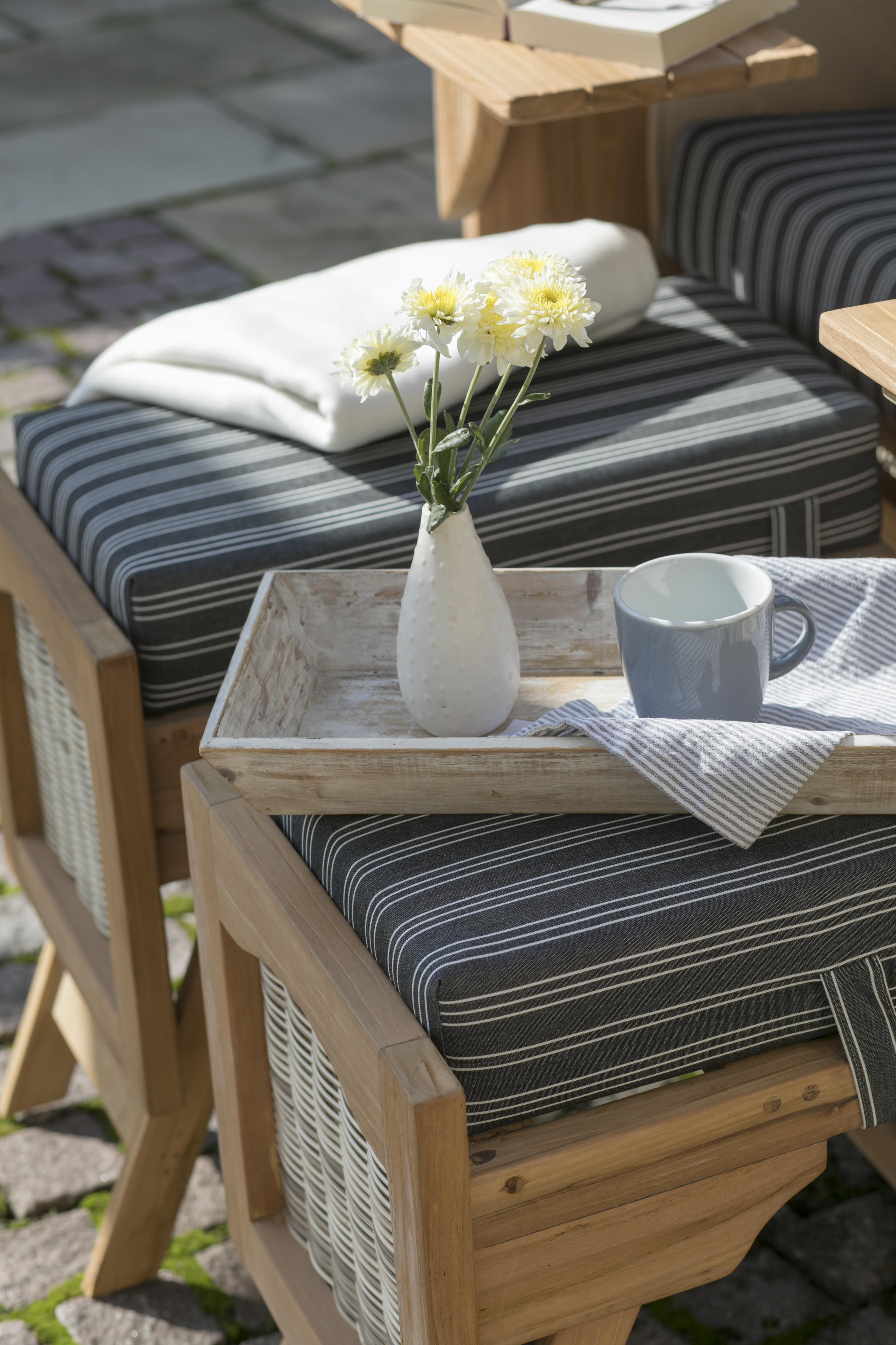 Ein Holztablett auf einem gestreiften Outdoor-Kissen hält eine weiße Vase mit gelben Blumen und einen Becher – perfekte Akzente für Ihren Strandkorb DÜNE Design grau mit Nadelstreifen inkl. Feuerschale gratis.