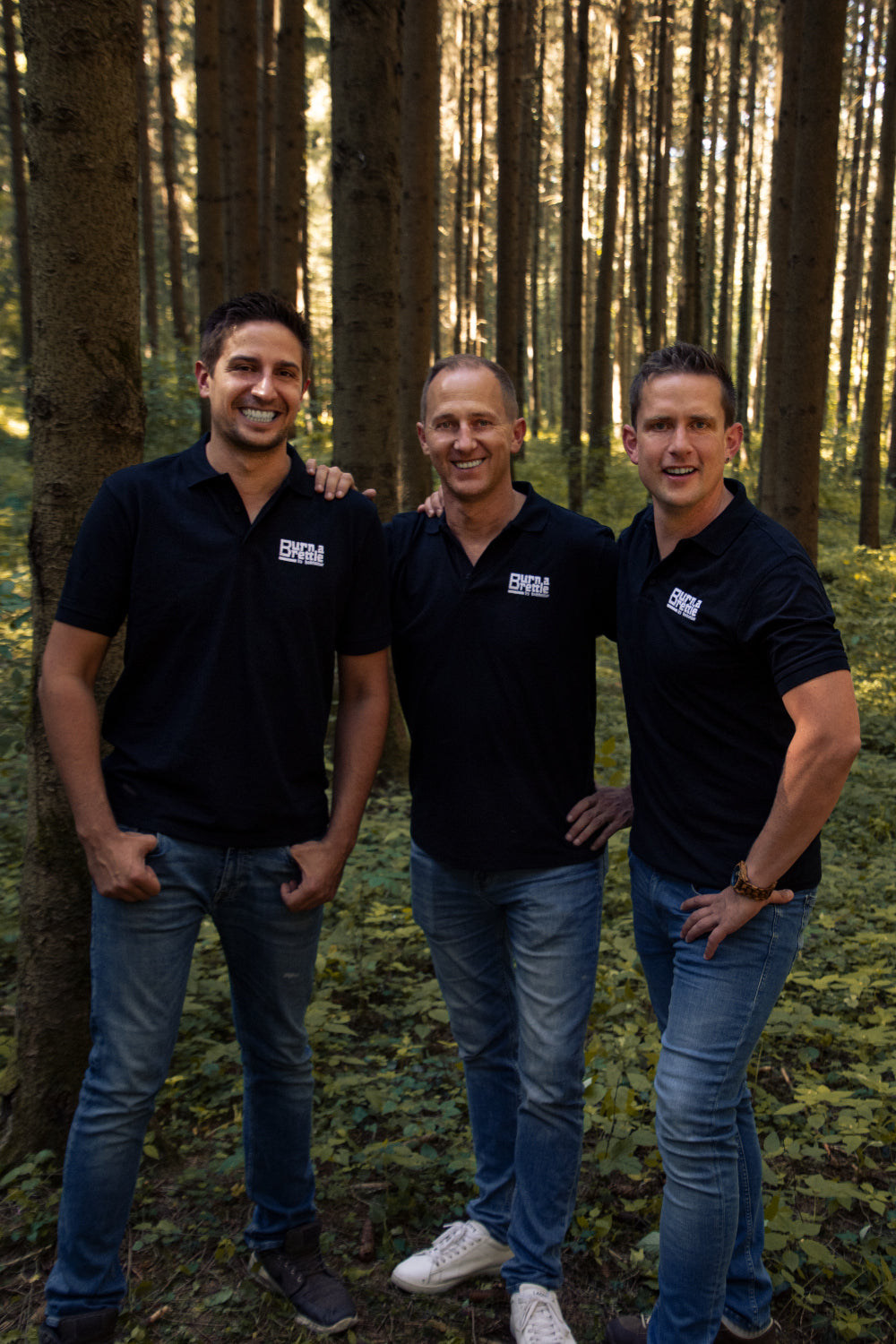 Drei Männer in schwarzen Poloshirts und Jeans stehen lächelnd zusammen in einem sonnenbeschienenen Wald und diskutieren vielleicht über eine „Burn a Brettle Grill“-Idee, die von nachhaltigen Materialien inspiriert ist.