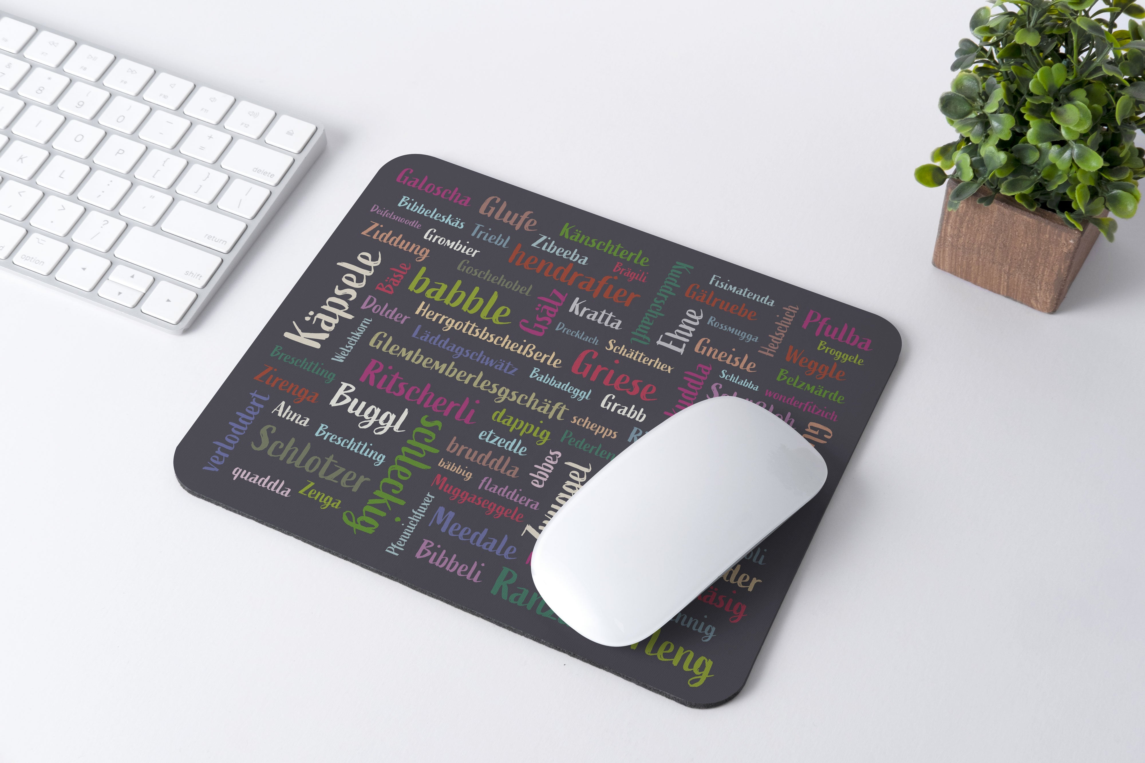 Auf einem Schreibtisch liegen eine weiße Maus und eine Tastatur mit dem Mundart-Mousepad, das ein stilvolles Wortwolken-Design für Oberflächenschutz aufweist, begleitet von einer kleinen Topfpflanze.
