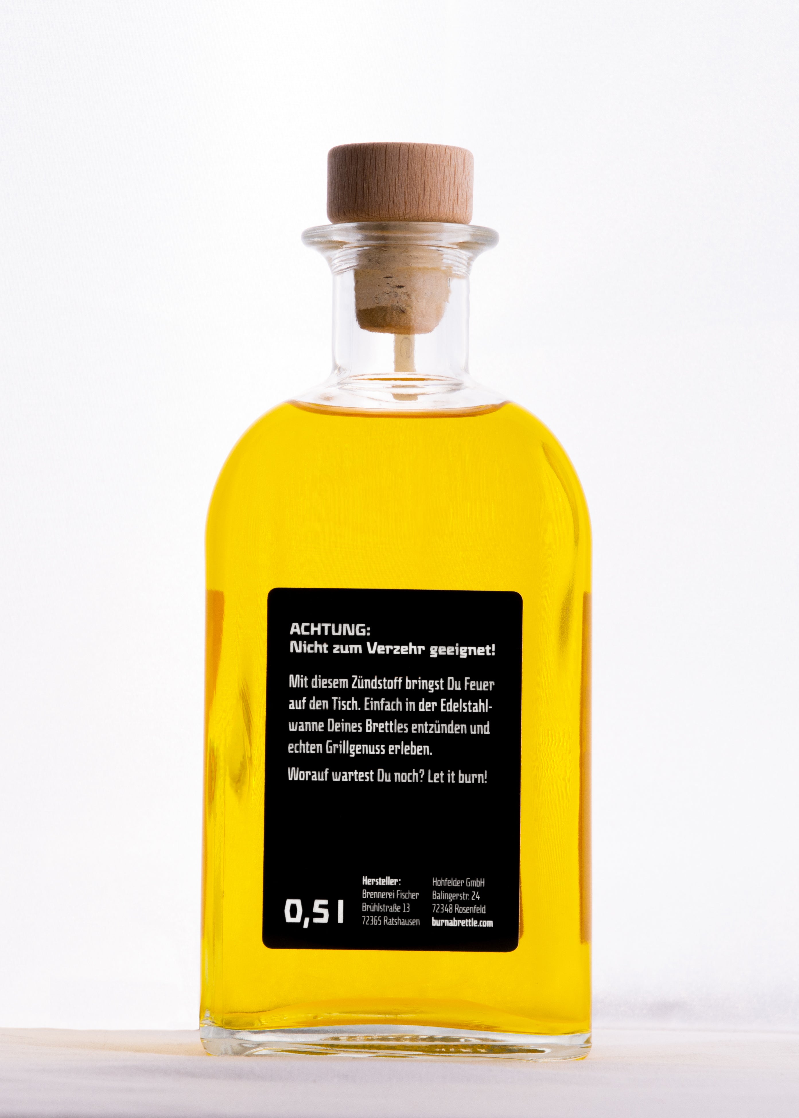 Glasflasche mit gelber Flüssigkeit, mit Holzstopfen und schwarzem Etikett mit deutschen Warnhinweisen und „Verbrenne einen Brettle Zündstoff“ auf weißem Hintergrund.