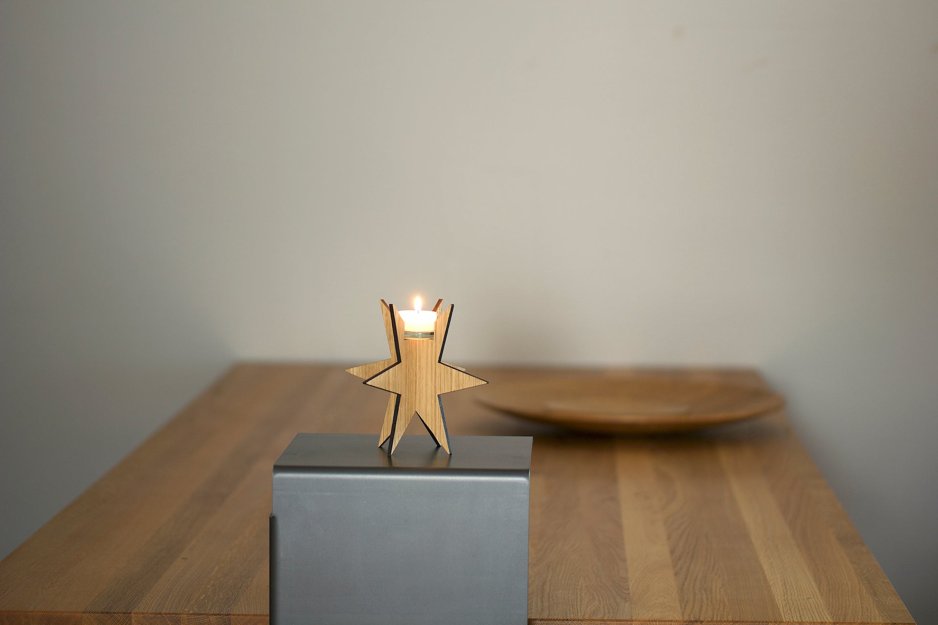 Eine brennende Kerze in einem Teelichthalter „Sternenlicht“ steht auf einem Tisch und verbreitet ein warmes Licht mit einem Holzteller im Hintergrund.