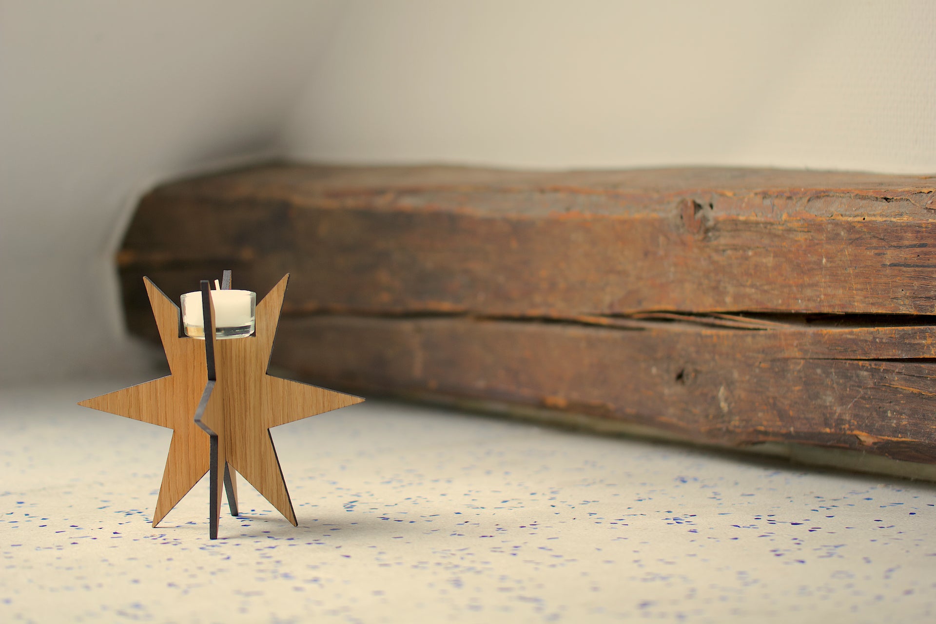 Der Teelichthalter Sternenlicht, aus Holz in Sternform mit weißem Kerzenlicht, dekorativ auf gesprenkelter Fläche neben einem Holzbalken platziert – ideal für deine Weihnachtstradition.