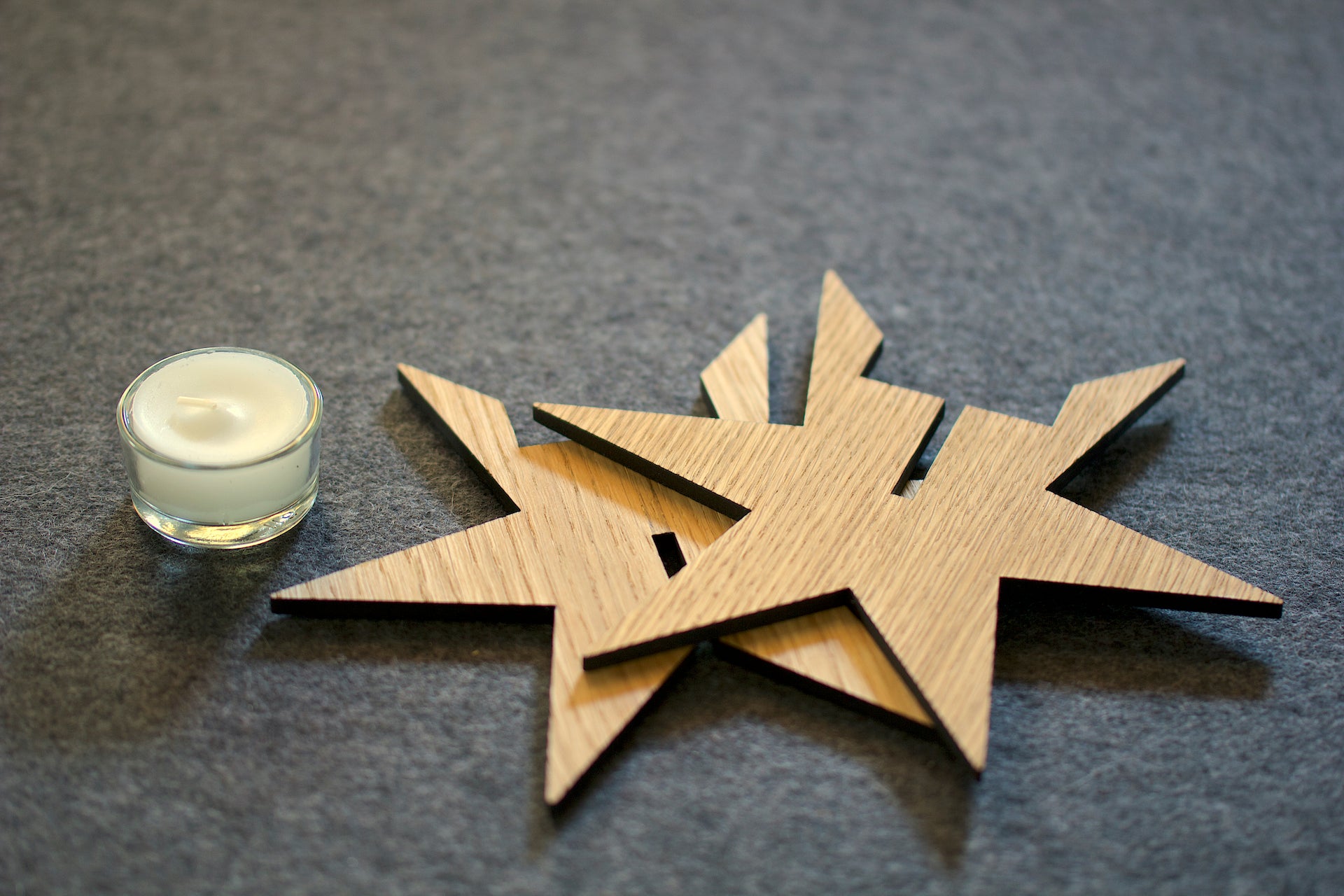 Eine kleine weiße Kerze in einem Teelichthalter Sternenlicht sitzt neben hölzernen Sternformen auf einer grauen Oberfläche und sorgt für eine gemütliche Weihnachtstradition.