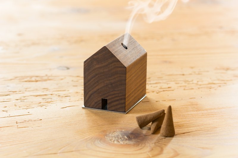 Das Räuchermännchen Rauchhaus hat die Form eines hölzernen Schwarzwaldhauses und stößt Rauch aus, während drei Räucherkegel auf einer nahe gelegenen Holzoberfläche ruhen.