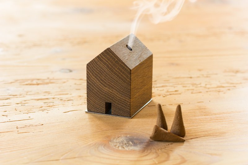 Ein Räuchermännchen-Rauchhaus – ein kleines Holzhaus, aus dessen Schornstein Rauch aufsteigt – steht neben zwei dreieckigen Räucherkegeln auf dem Tisch und sorgt für eine bezaubernde Szene.