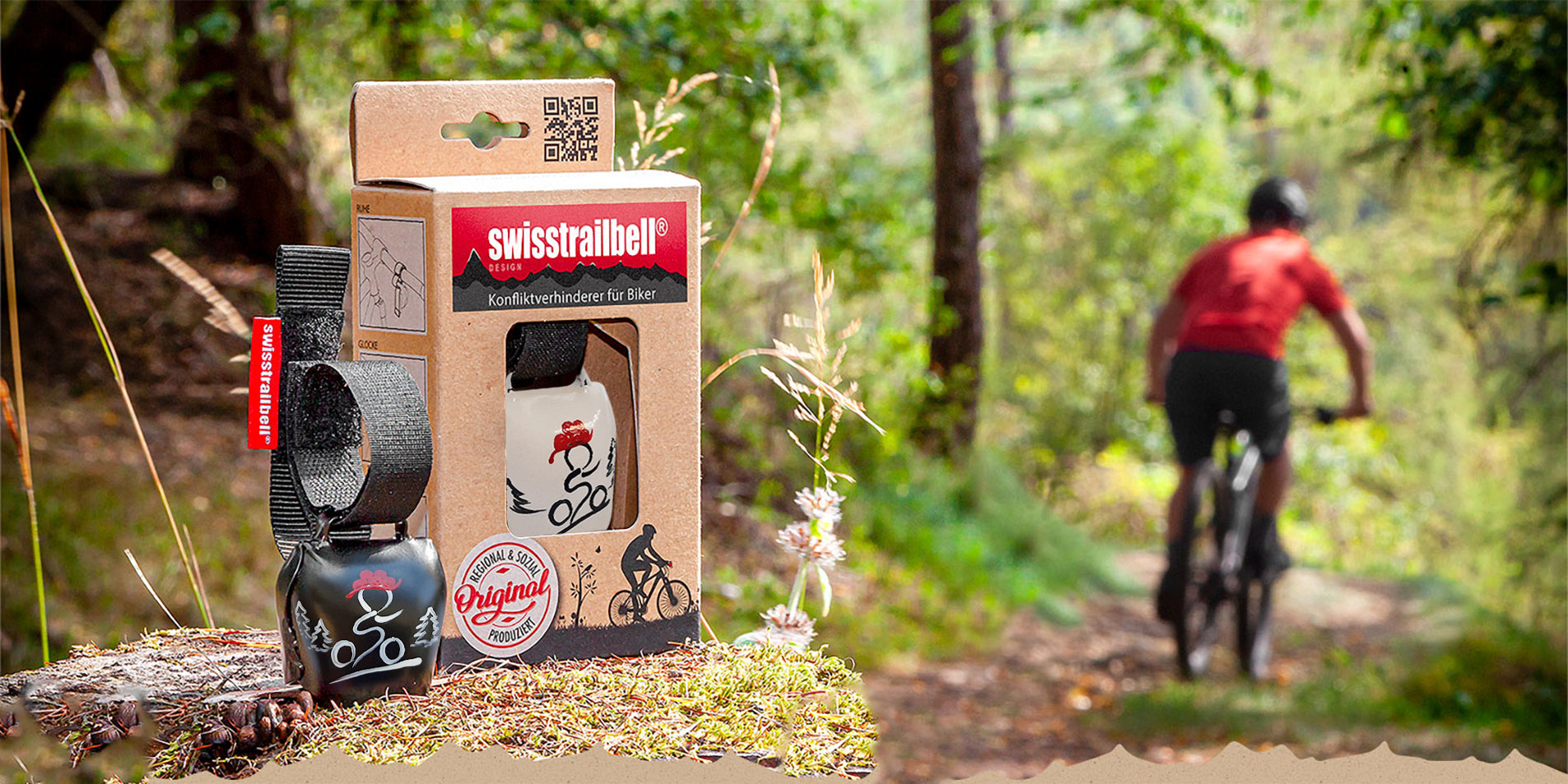 Eine verpackte Fahrradglocke swisstrailbell® steht auf einem Baumstumpf, während im Hintergrund ein Radfahrer einen Waldweg entlang fährt.