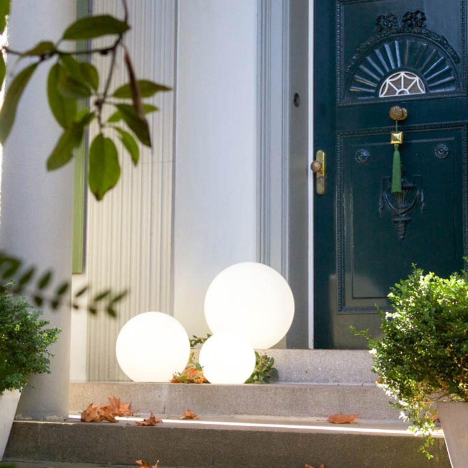Drei leuchtend weiße Leuchtkugeln und Solarlichter schmücken die Stufen einer dunkelgrünen Tür, umgeben von Pflanzen und Herbstblättern, und schaffen eine zauberhafte Gartenszene, die von diesen eleganten Solar-Gartenlichtern beleuchtet wird.
