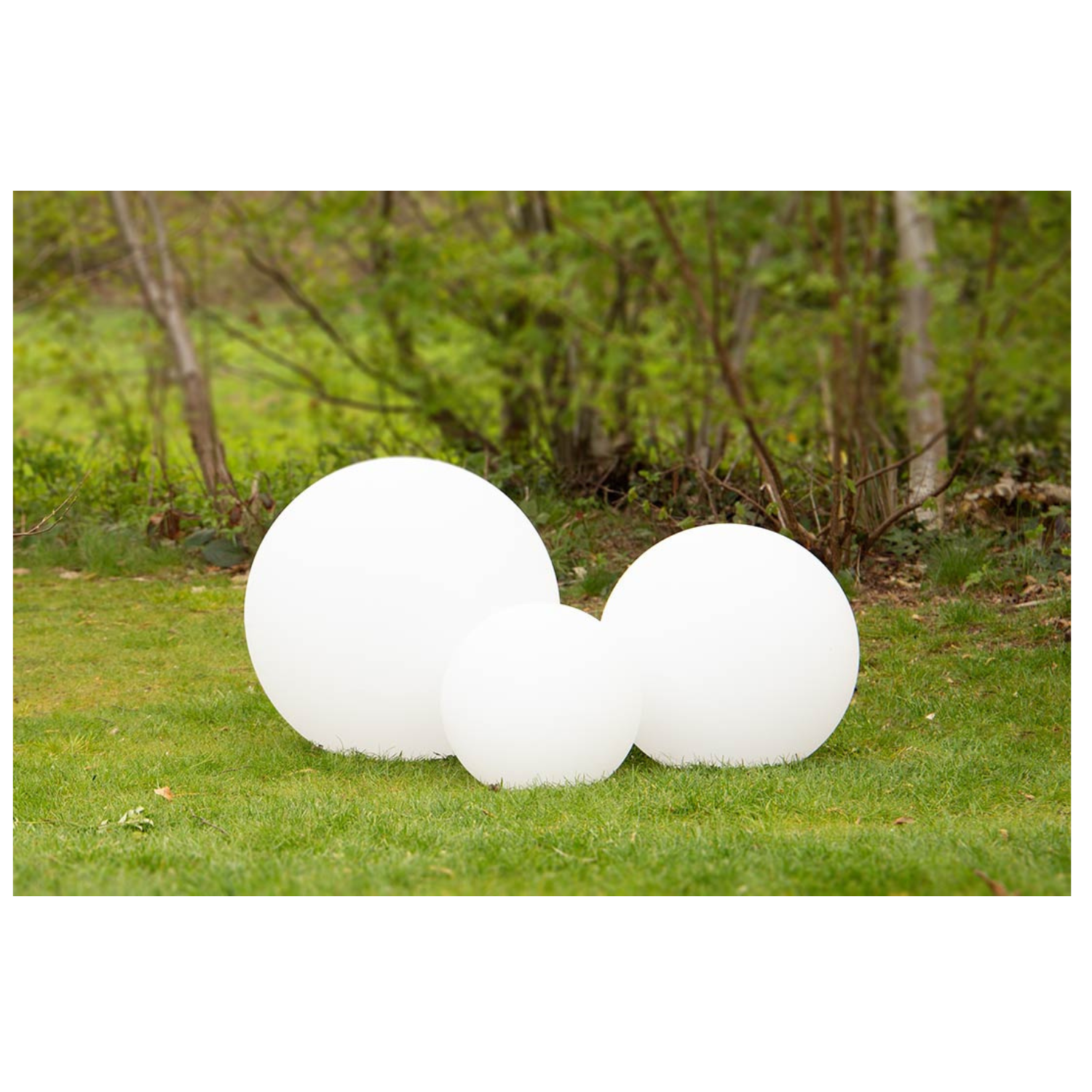 Drei große, weiß leuchtende Kugeln, darunter die Leuchtkugel Solar, stehen auf grünem Gras mit Bäumen im Hintergrund.