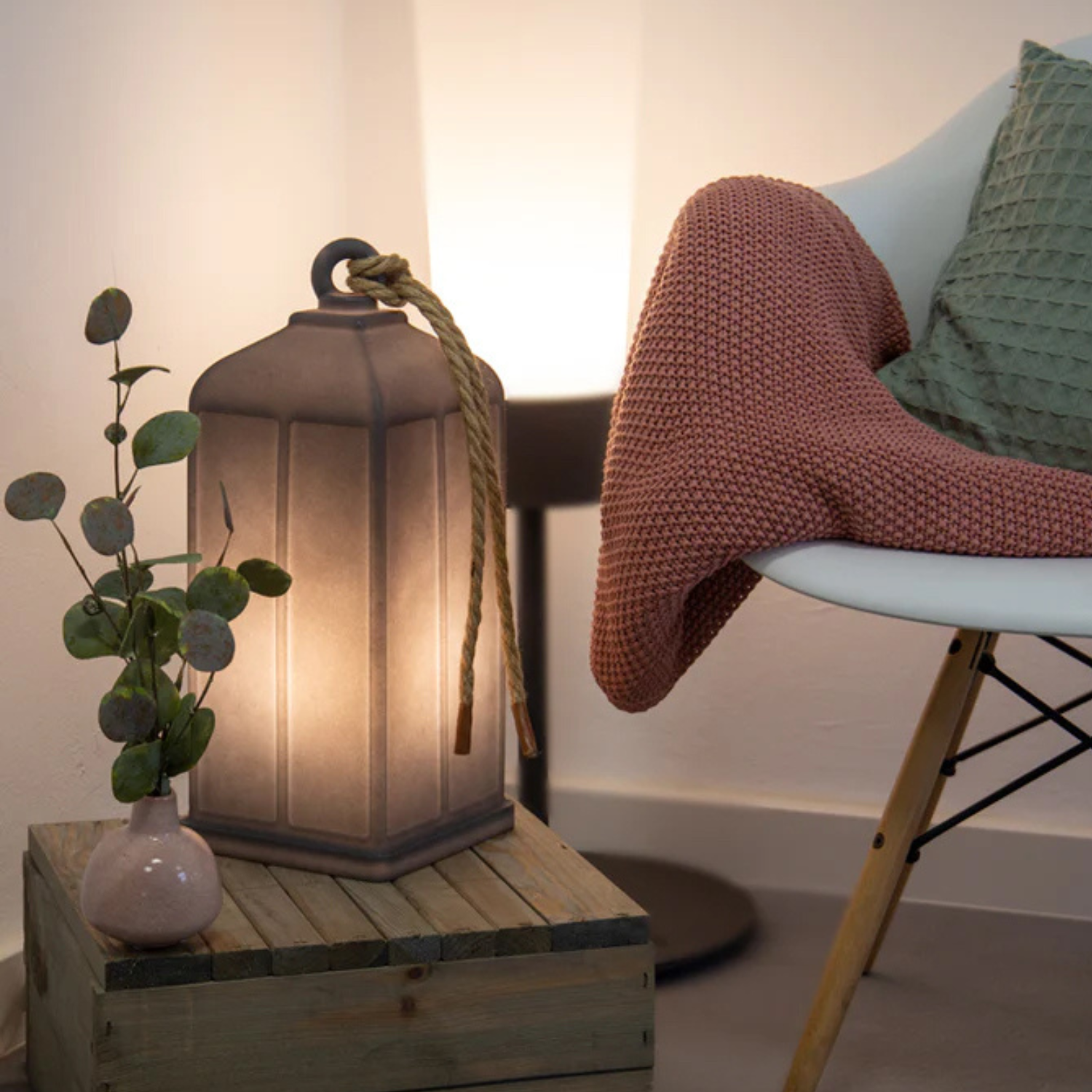 Gemütliche Ecke mit Sessel, grünem Kissen, rosafarbener Decke, leuchtender Laterne, Solar mit Dämmerungssensor, Vase mit Grünpflanzen und einer Holzkiste.