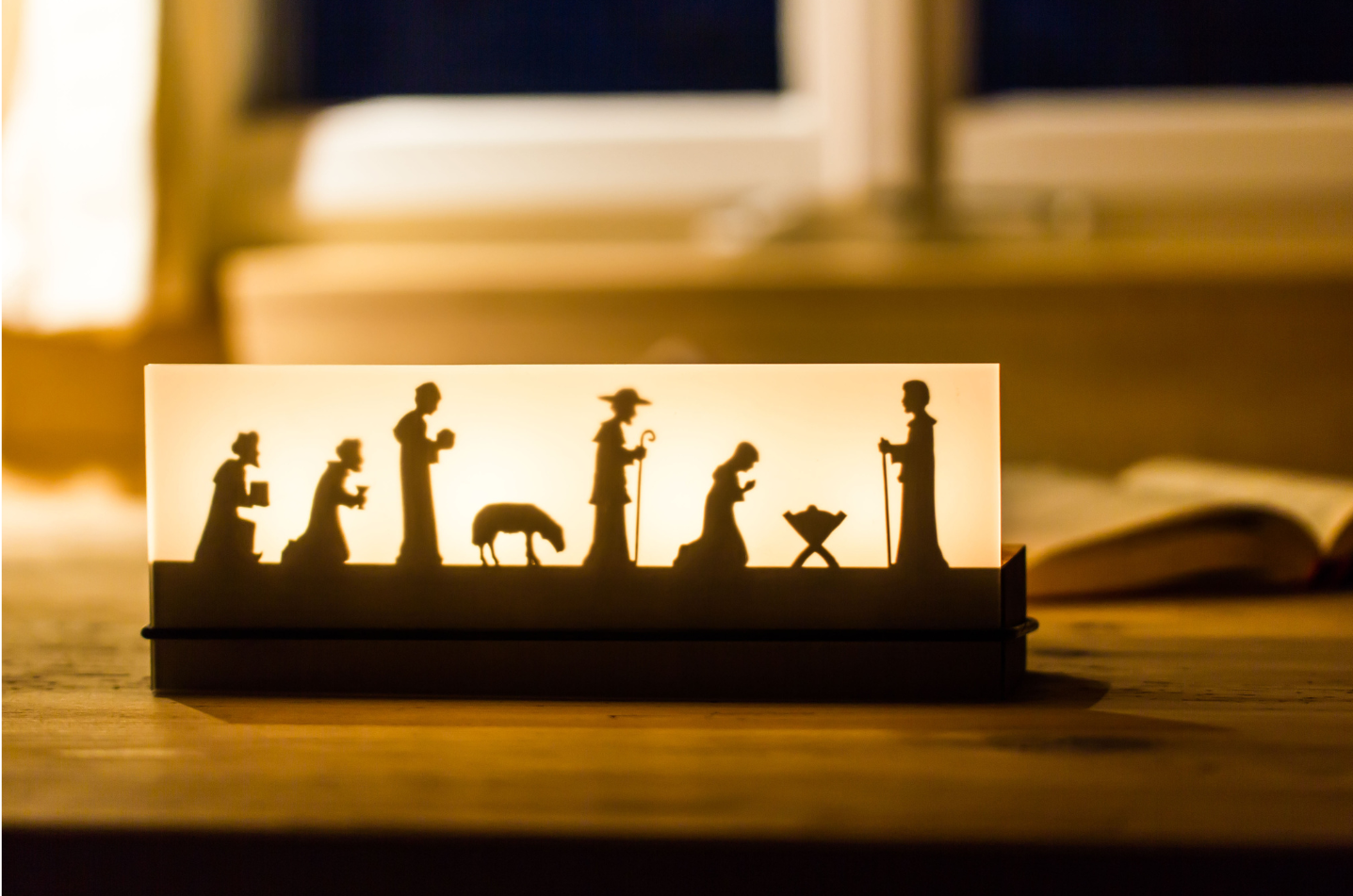 Der Teelichtleuchter Schattenspiel 4er Krippe beleuchtet eine Krippensilhouette auf einem Holztisch am Fenster und stellt so die Weihnachtsgeschichte wunderschön dar.