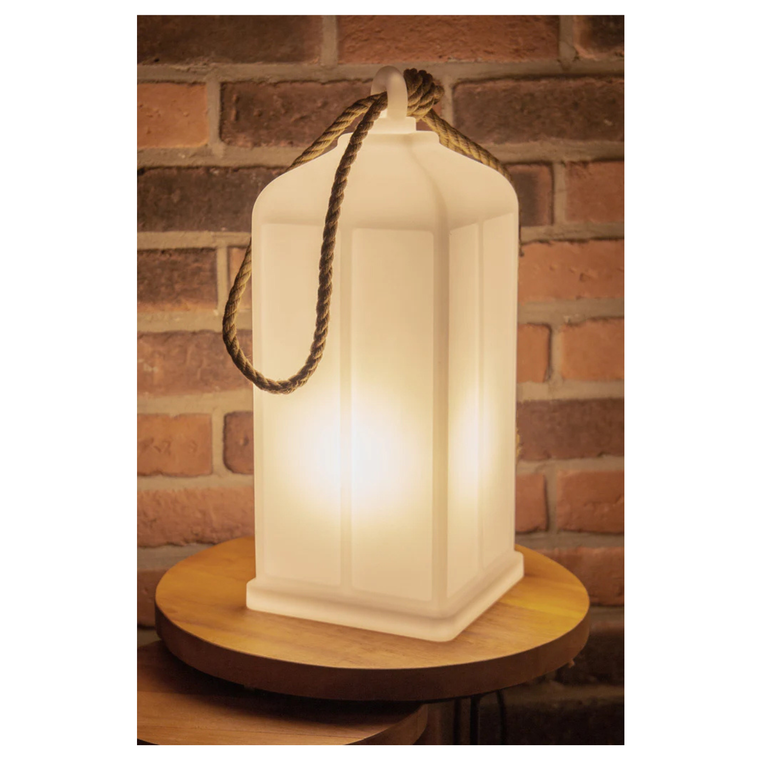 Die leuchtende Laterne, Solar mit Dämmerungssensor und Seilgriff steht wetterfest auf einem runden Holztisch vor einer Backsteinwand.