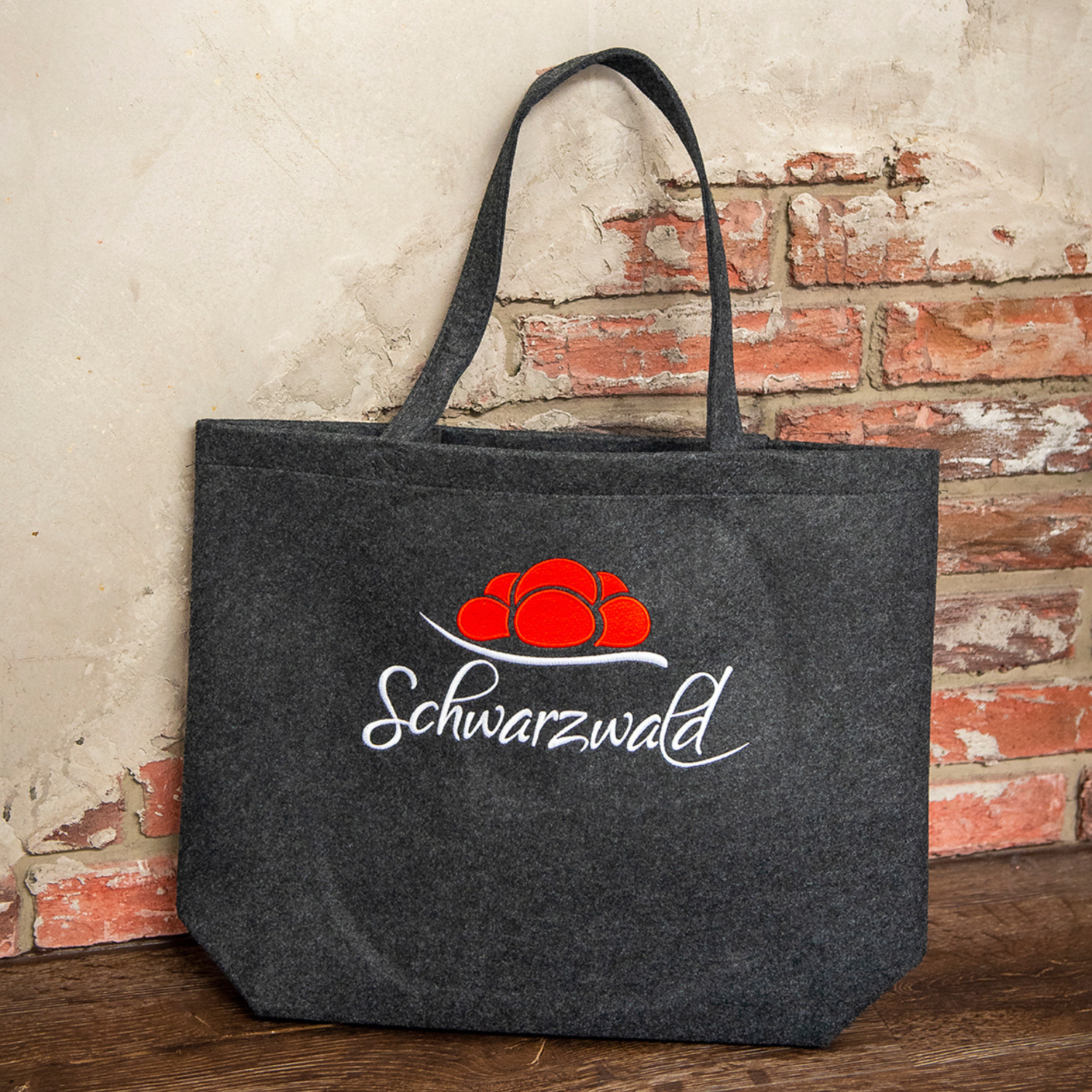 Der Filz-Shopper - Schwarzwald ist eine graue Filztasche mit Schwarzwald-Motiv und roter Mützengrafik, versehen mit raffinierter Stickerei, präsentiert vor einer rustikalen Backsteinmauer.
