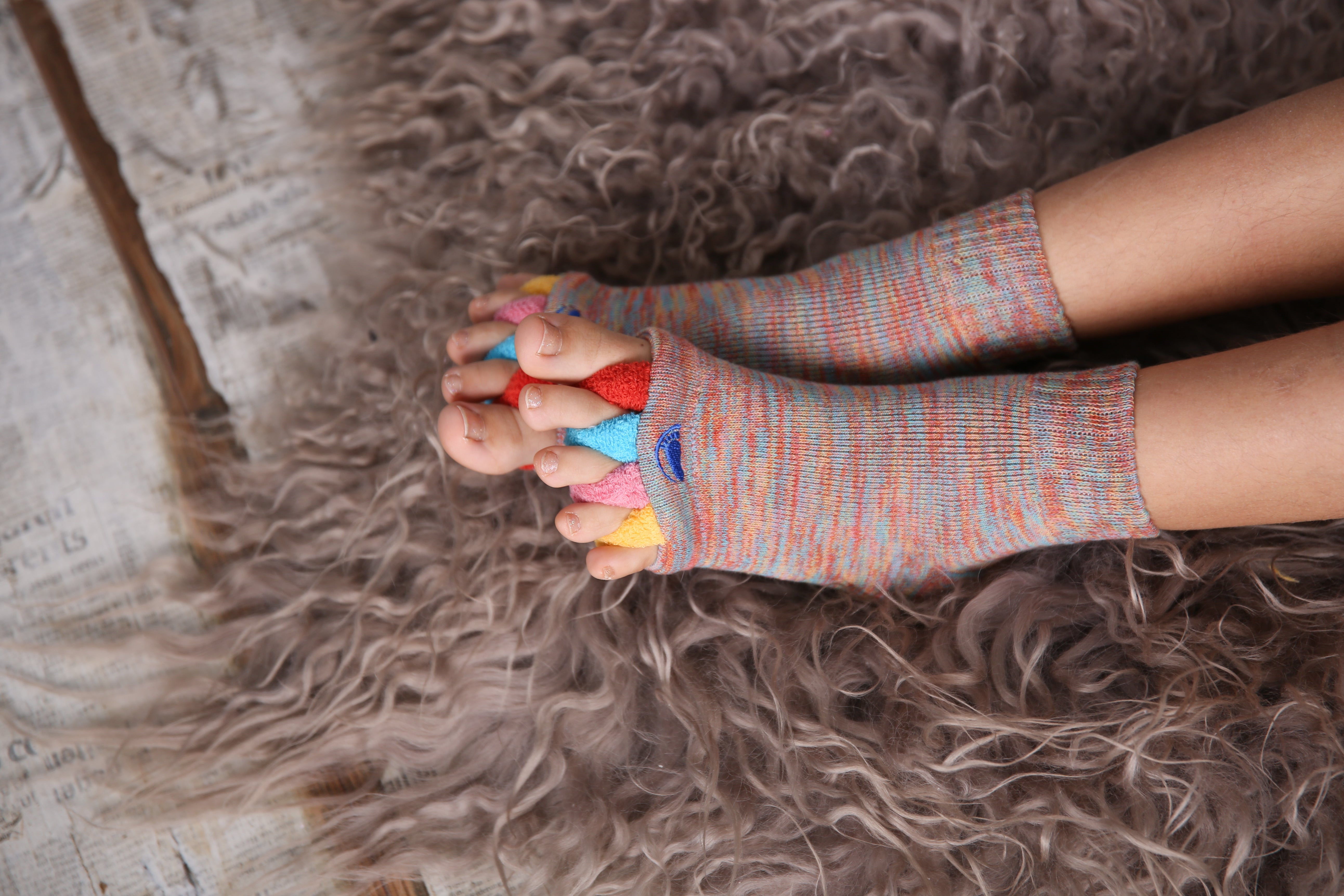 Ein Kind sitzt auf einem flauschigen Teppich, die Zehen schauen durch die Öffnungen der Fußausrichtungssocken „Happy Feet“ – diese farbenfrohen Socken bieten spielerischen Komfort und sind ein therapeutisches Hilfsmittel bei Fußbeschwerden.