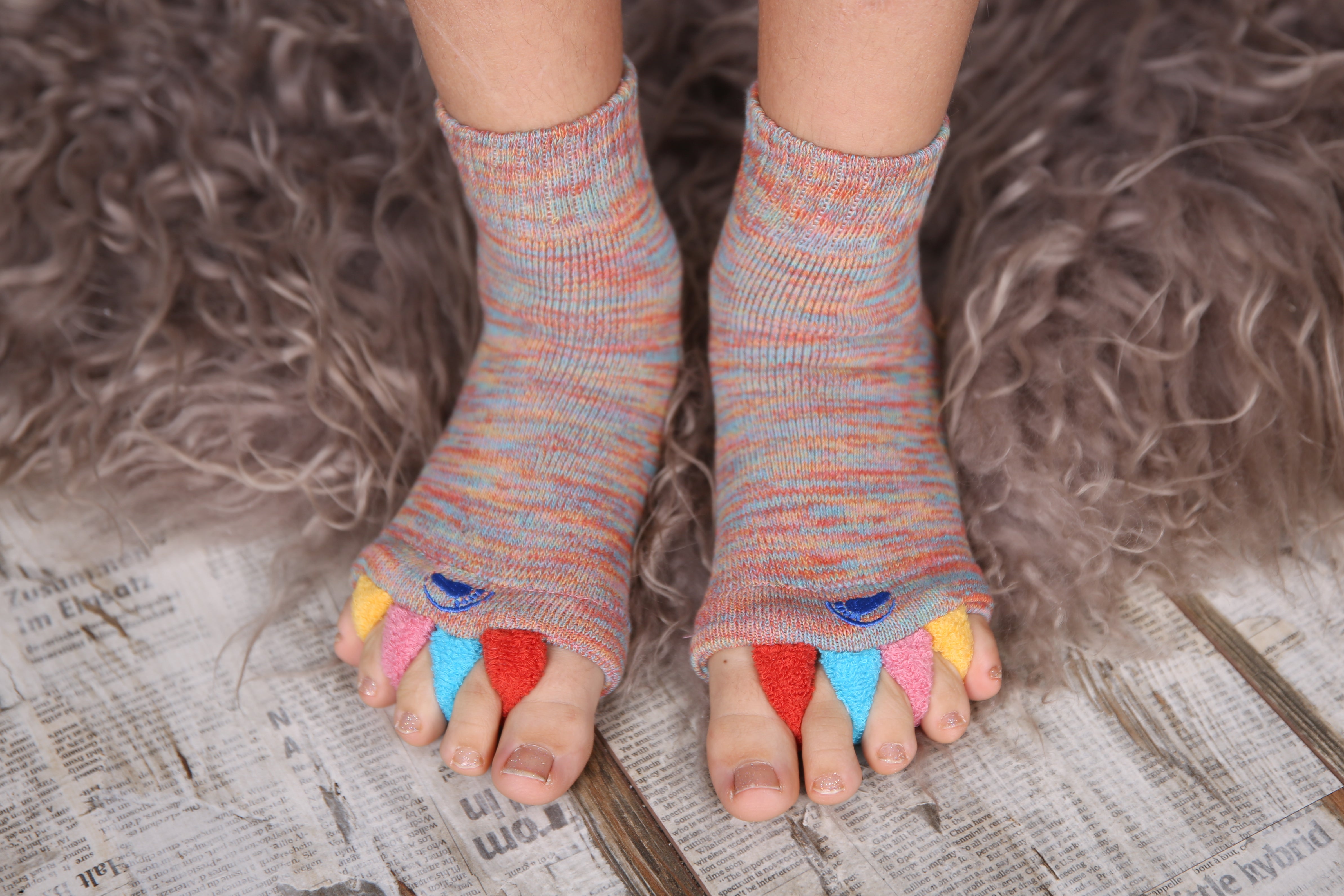 Bunte Fußausrichtungssocken „Happy Feet“ ruhen auf einem pelzigen Teppich über Holz- und Zeitungsmusterböden – ein ideales, cleveres therapeutisches Hilfsmittel zur Linderung von Fußbeschwerden.
