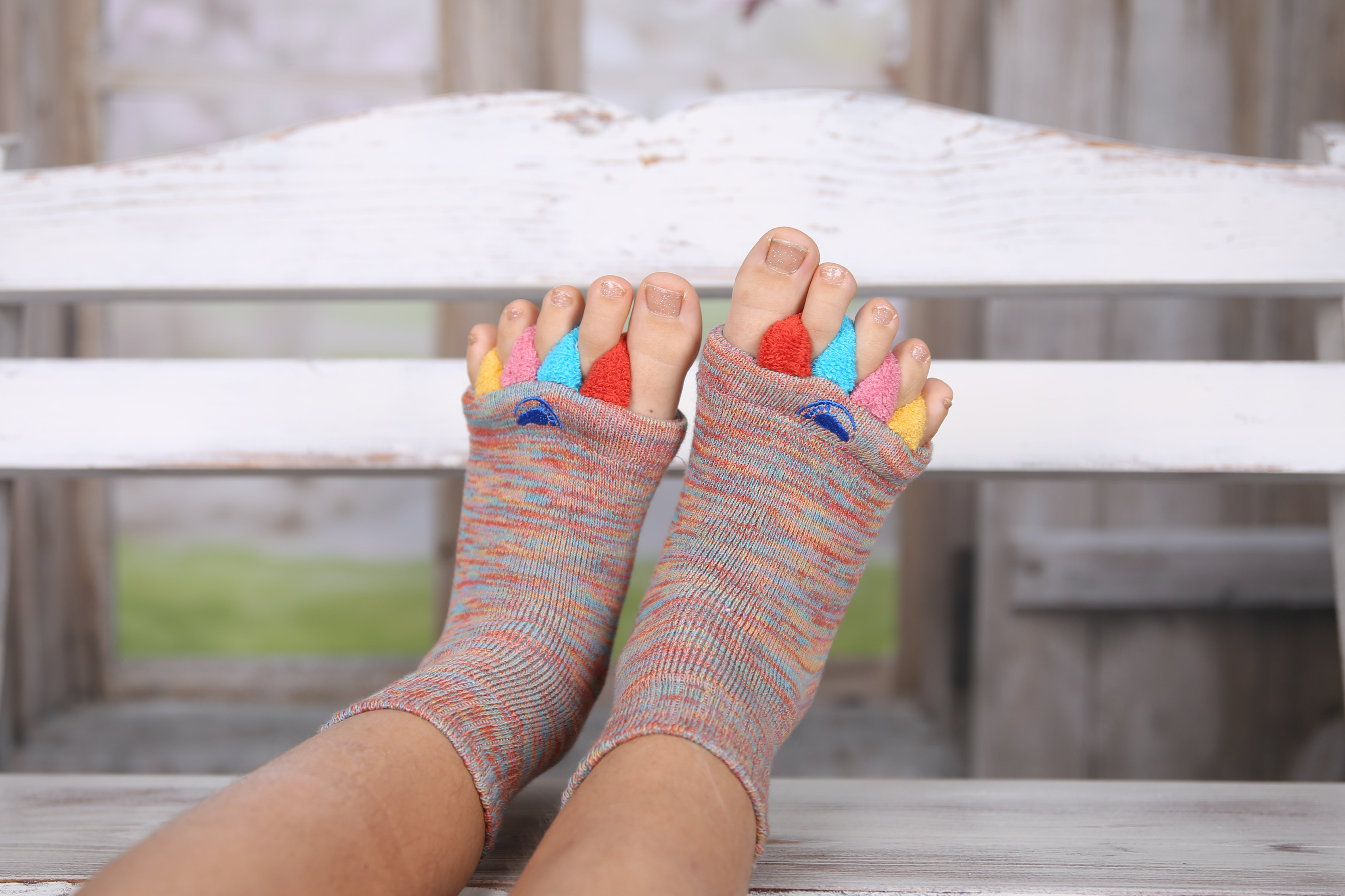 Person in den offenen Fußausrichtungssocken „Happy Feet“ mit bunten Zehentrennern sitzt auf einer weißen Holzbank – ideal als therapeutisches Hilfsmittel bei Fußbeschwerden.