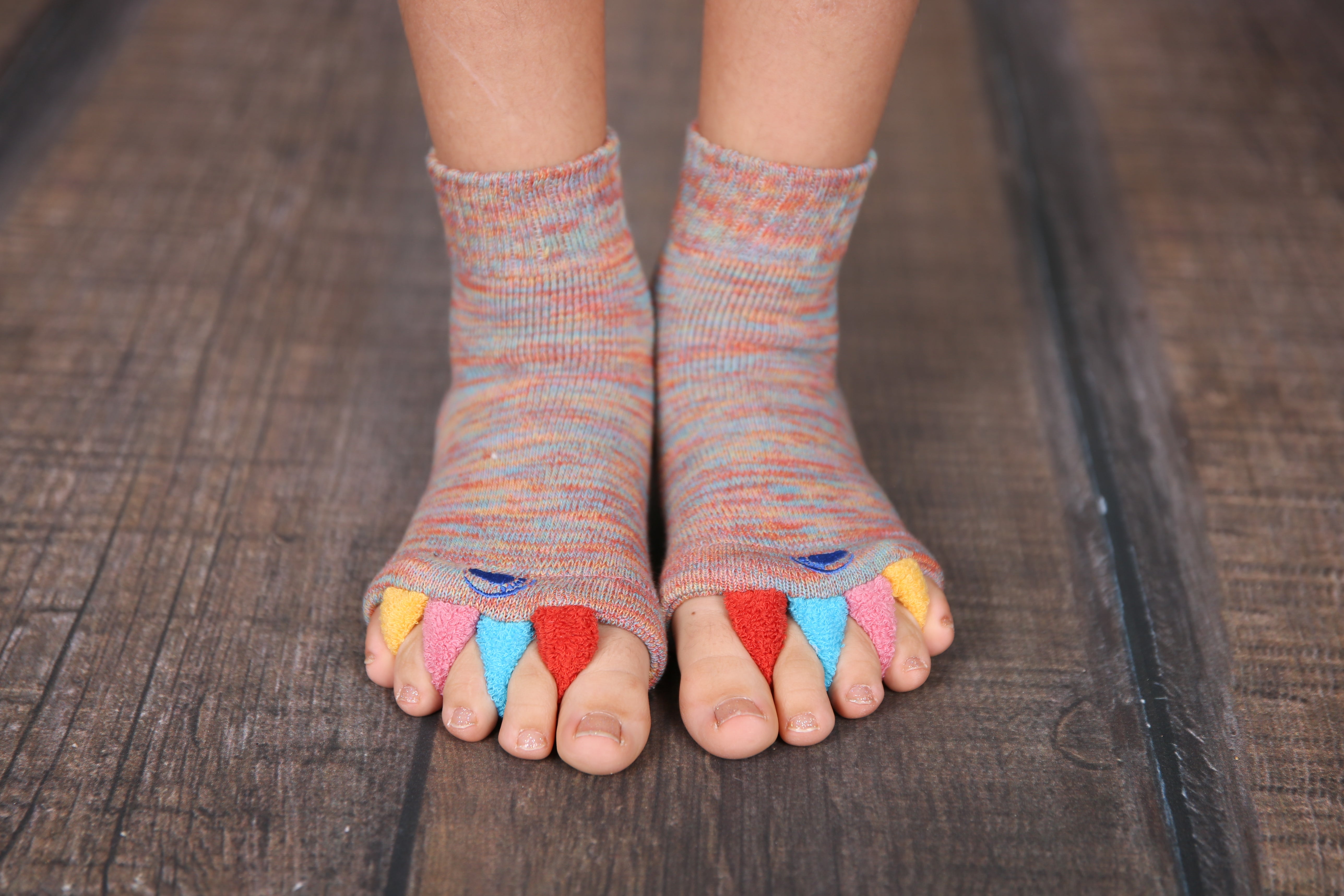 Ein Kind trägt die Fußausrichtungssocken „Happy Feet“, bei denen jeder Zeh eine andere Farbe hat und die Enden offen sind, und steht auf einem Holzboden – ein spielerisches und zugleich therapeutisches Hilfsmittel, das Fußproblemen vorbeugen soll.